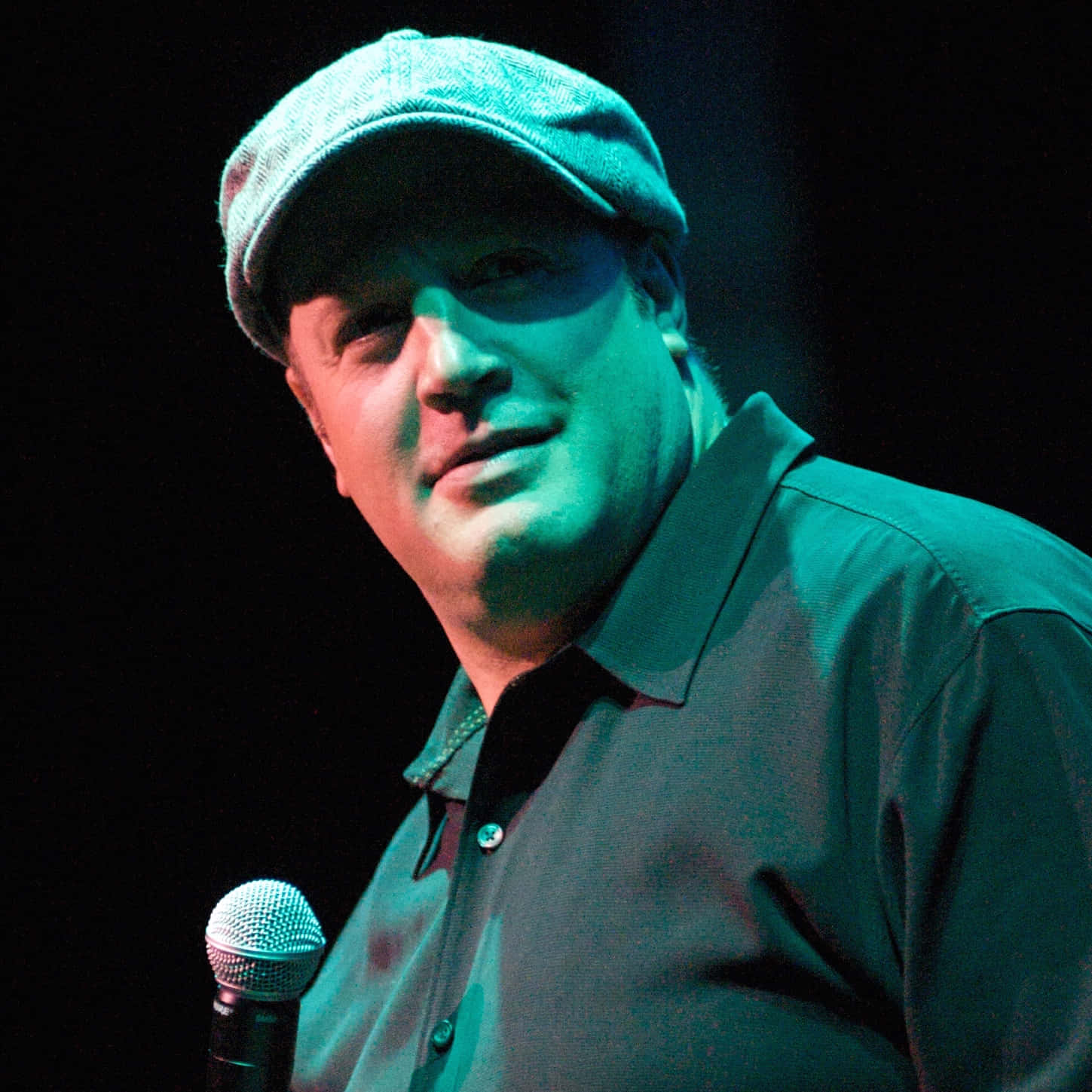 Kevin James 桌布 壁紙 — 一名戴帽及穿有領襯衫的男子在綠色舞台燈光下微笑著手持麥克風。