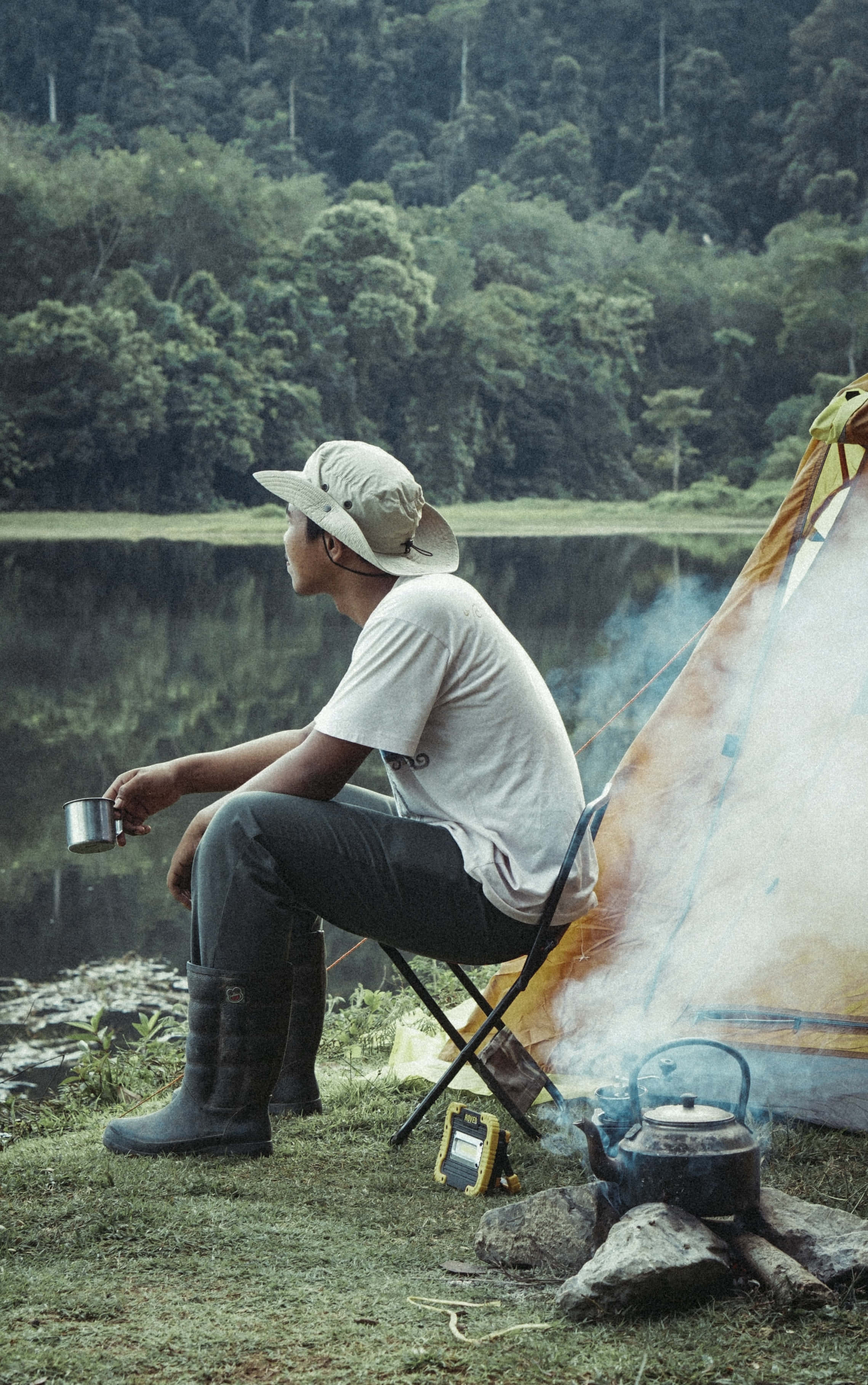 Camping 桌布 壁紙 — 一名戴帽子的男人坐在湖邊營火旁，旁邊有一個帳篷，他正從杯子裡小口喝著，煙霧向上繚繞。