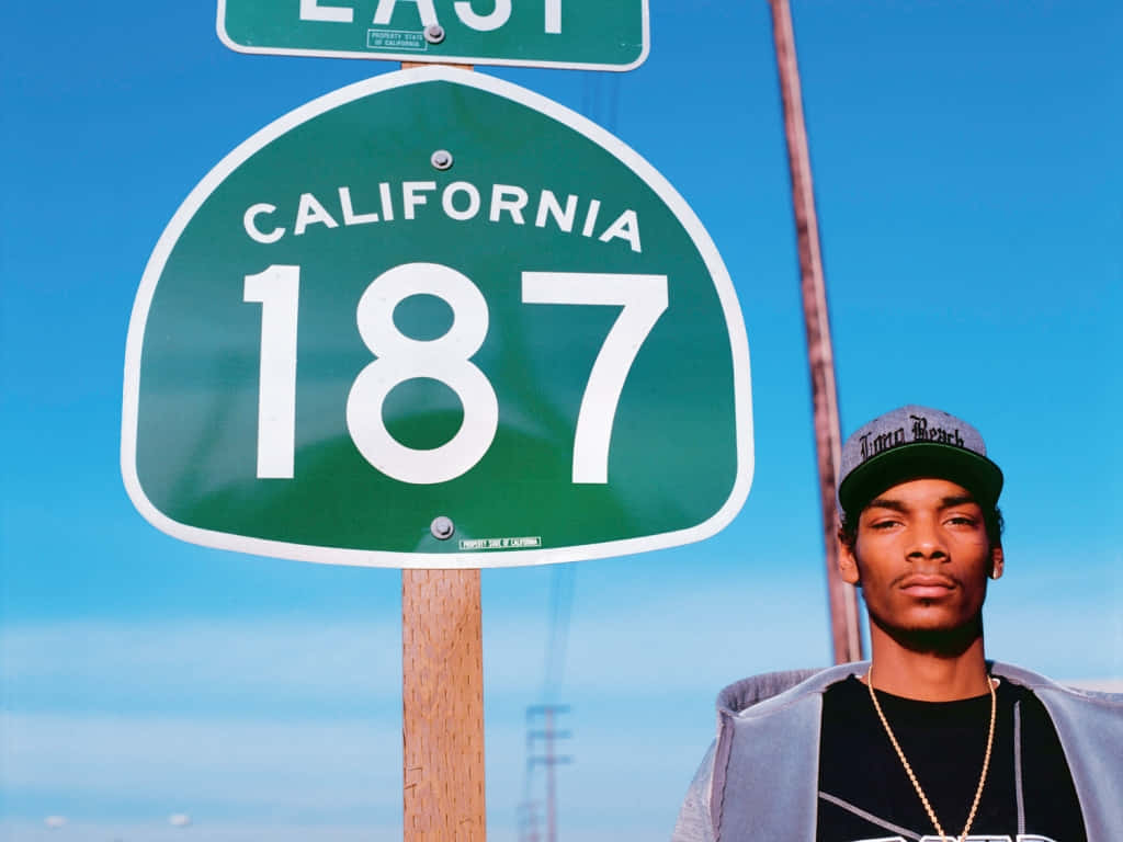 Young Snoop Dogg 桌布 壁紙 — 一名戴帽子的男子站在綠色加州187號公路標誌旁，天空湛藍清澈。