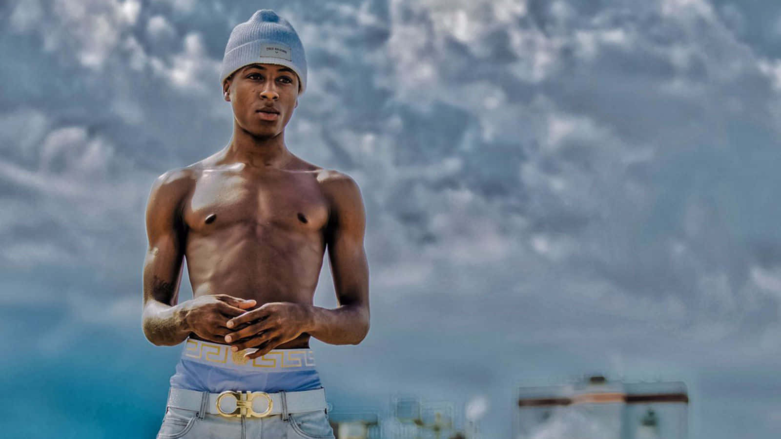Youngboy 桌布 壁紙 — 一名戴毛帽的年輕男子赤裸上身，自信地站在戲劇性的多雲天空前，背後有模糊的城市建築。