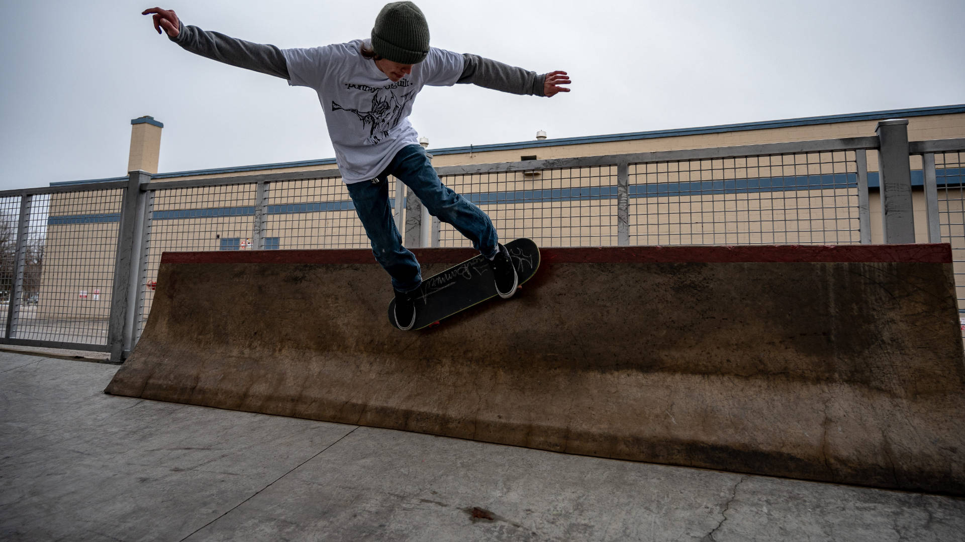 Skater Boy 桌布 壁紙 — 一名戴毛帽穿牛仔褲的滑板手，在陰天之下於一條風化的混凝土斜坡上磨滑。