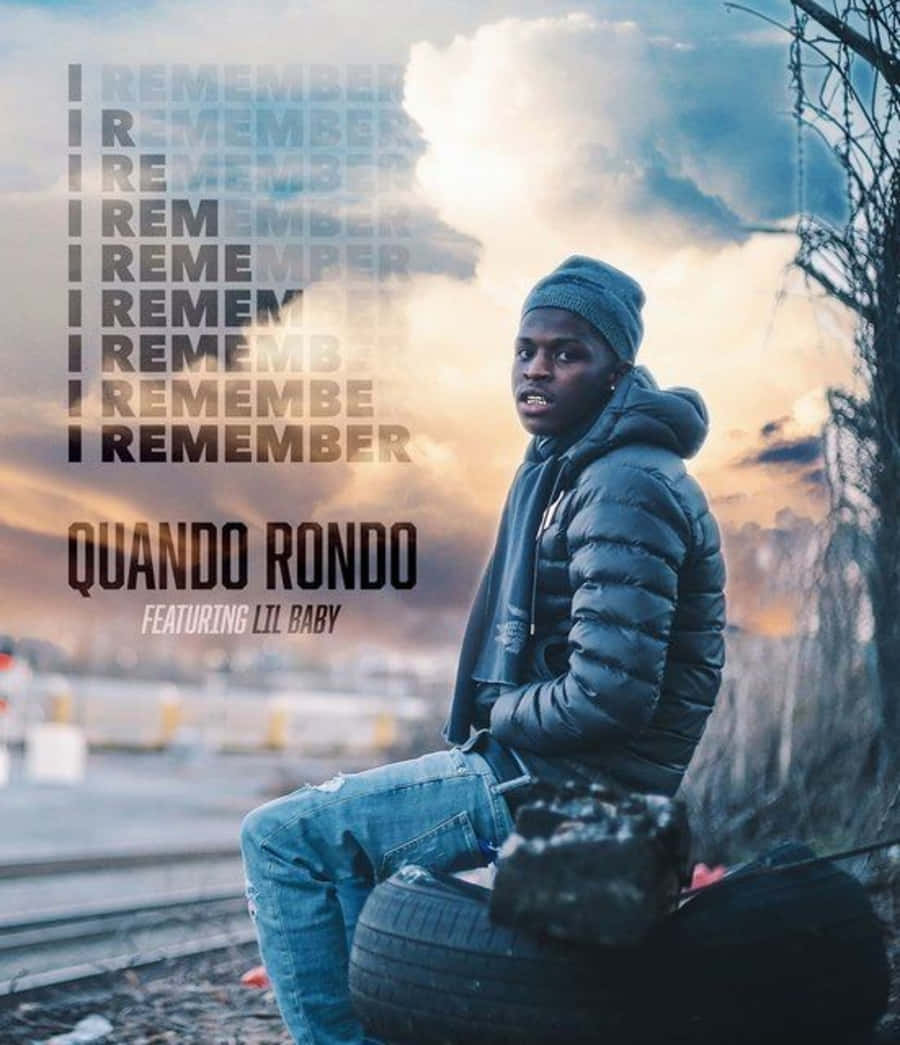 Quando Rondo 桌布 壁紙 — 一名戴毛帽穿羽絨外套的年輕男子坐在火車鐵軌旁的輪胎上，背景是戲劇性的日落天空，並有重複的文字疊加。