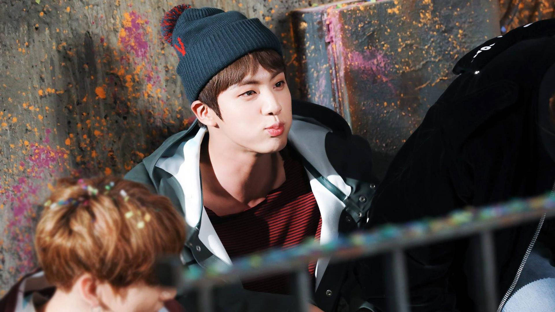 kim seok jin 桌布 壁紙 — 一名戴深色毛帽和穿外套的年輕男子對著佈滿塗鴉的牆壁做出鬼臉。