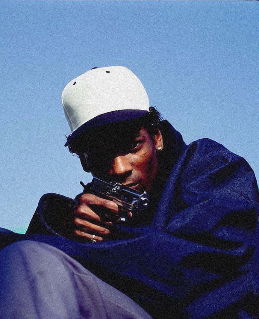 Young Snoop Dogg 桌布 壁紙 — 一名戴白帽、穿藍色外套的男子，在晴朗的藍天下專注地瞄準手槍。