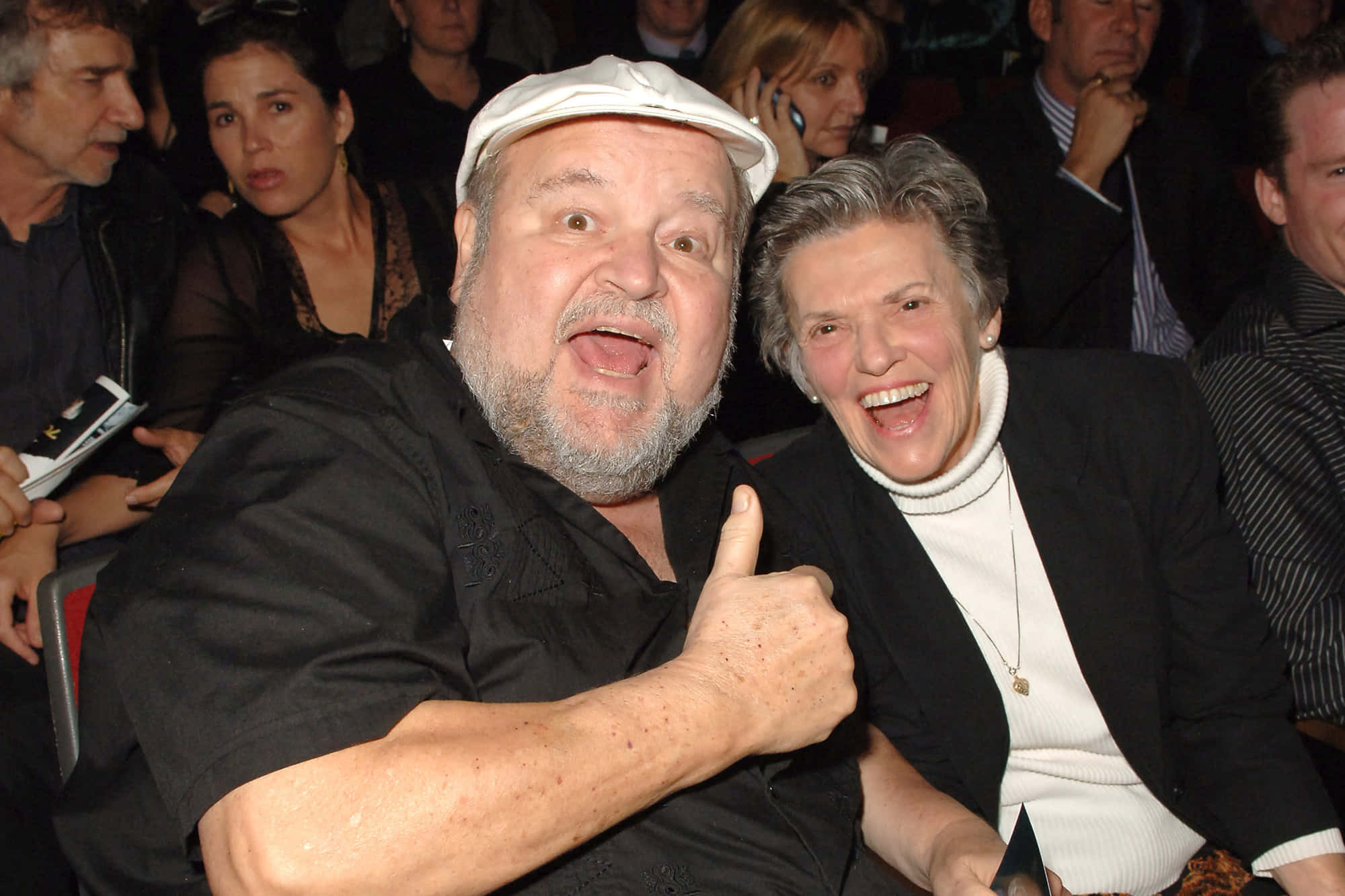 Dom Deluise 桌布 壁紙 — 一名戴白色帽子、穿黑色襯衫的男子在擁擠的活動中，豎起大拇指大笑，旁邊是一位穿白色高領毛衣的女子。
