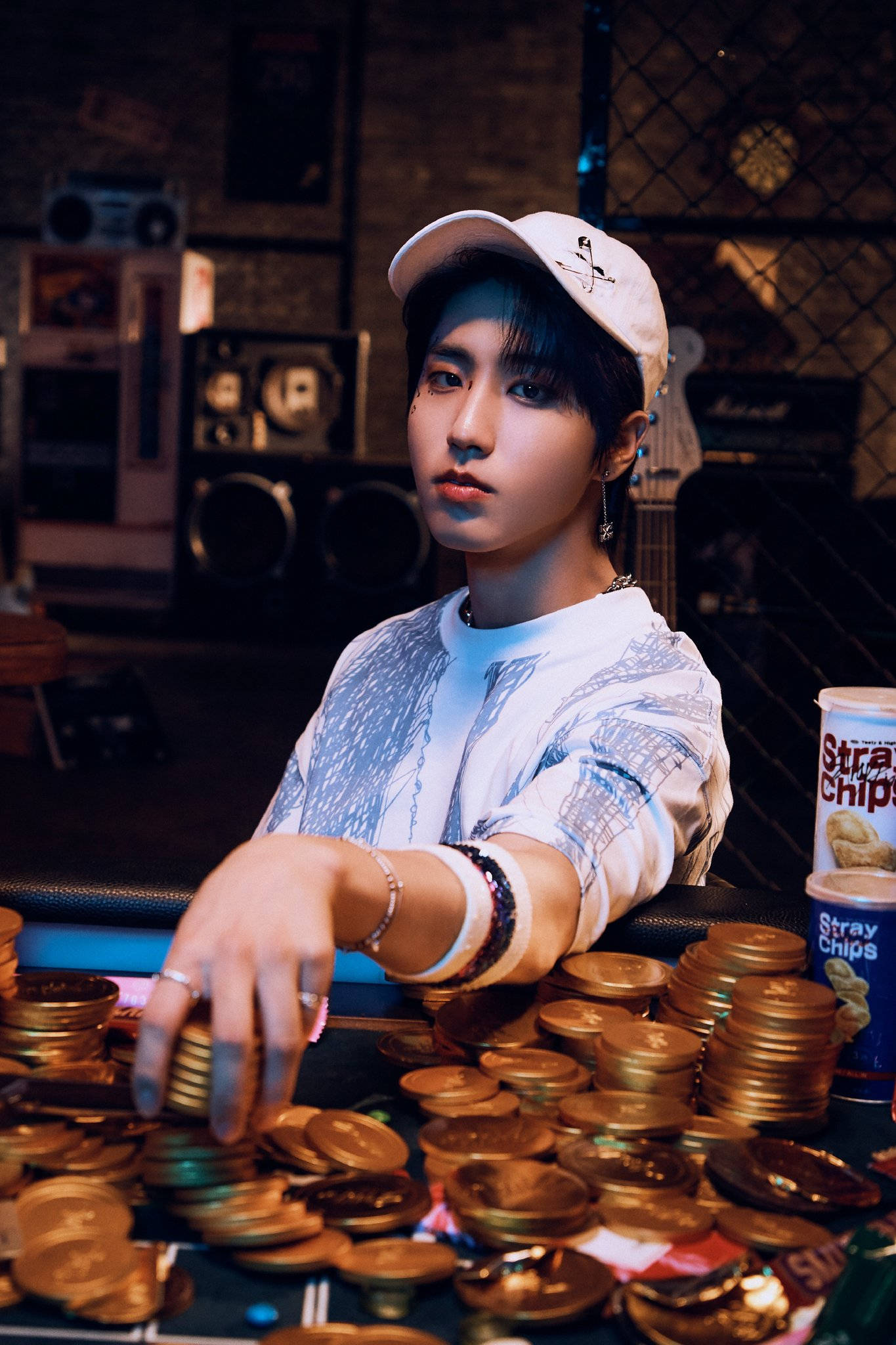 Stray Kids 桌布 壁紙 — 一名戴白色帽子和圖案襯衫的年輕男子坐在一張堆滿金幣和小吃罐的桌子旁，房間光線昏暗，風格復古。