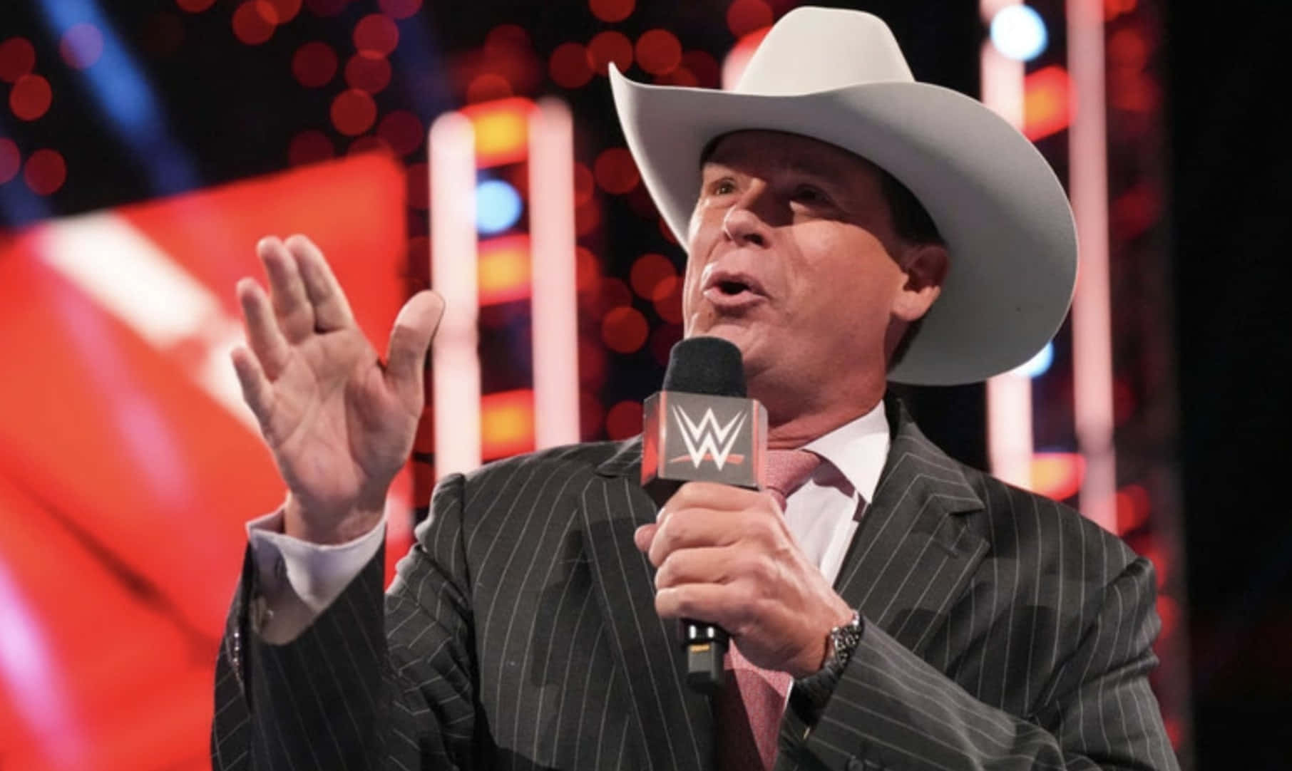 WWE 桌布 壁紙 — 一名戴白色牛仔帽、身穿細條紋西裝的男子，在燈光明亮的舞台上熱情地做手勢，手持WWE麥克風。
