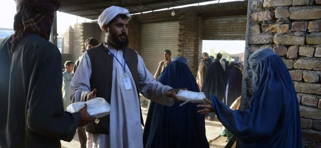 Taliban 桌布 壁紙 — 一名戴白色頭巾的男子在陽光照耀的磚砌庭院中，將食物包裹遞給一名穿藍色罩袍的女子。