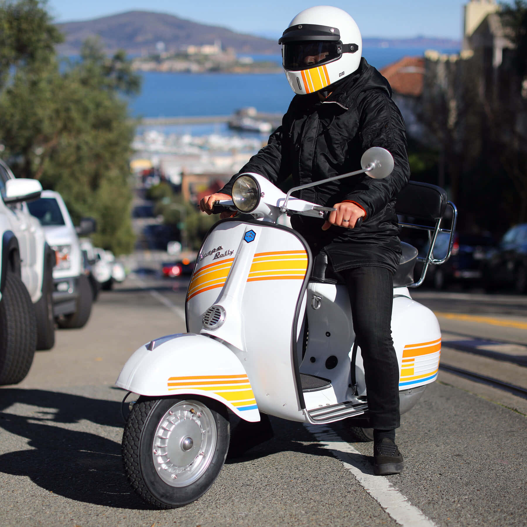 scooter 桌布 壁紙 — 一名戴白色頭盔、穿黑色外套的騎士，駕駛一輛帶有黃色條紋的白色Vespa Rally機車，沿著一條以海洋和山丘為背景的海岸街道行駛。