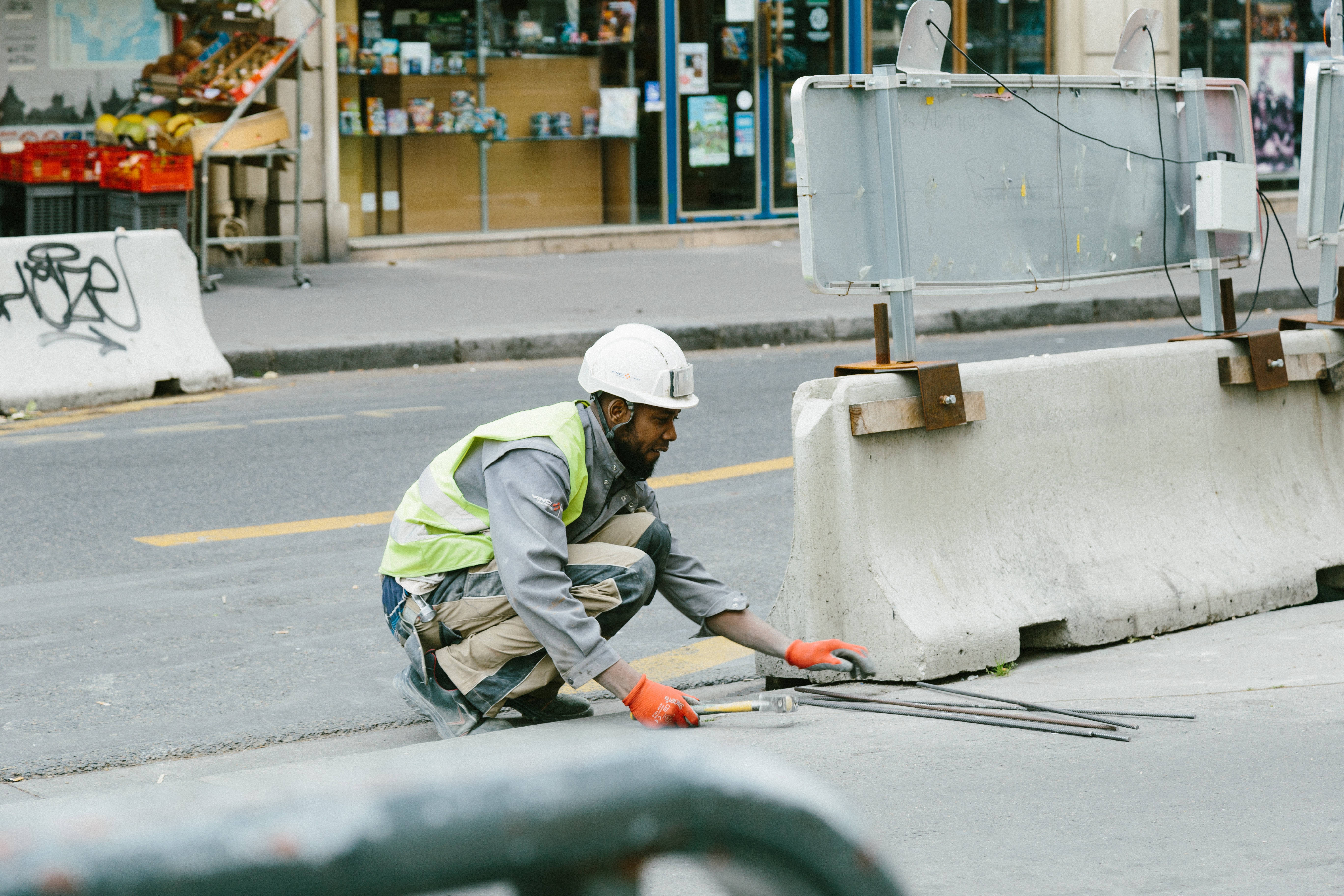 highway 桌布 壁紙 — 一名戴白色頭盔和黃色背心的建築工人跪在城市的街道上，在混凝土屏障附近用工具修補路面。