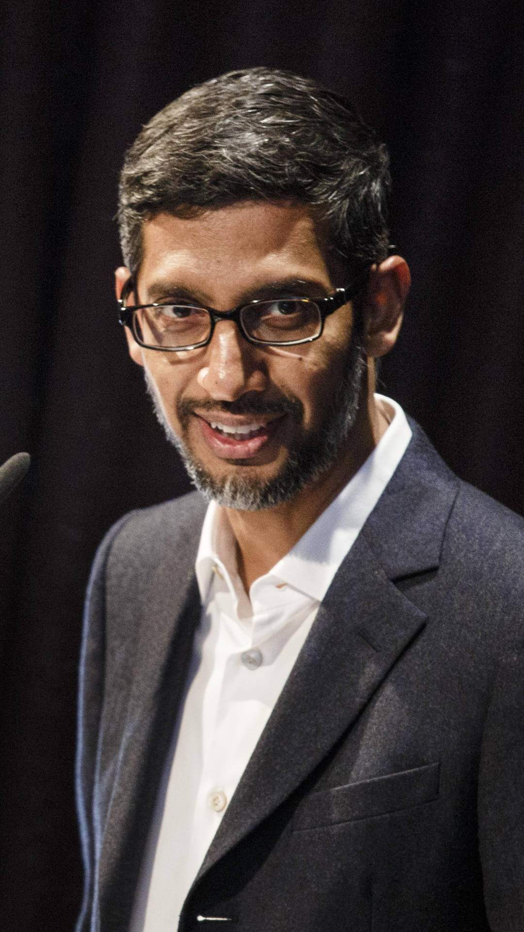 google 桌布 壁紙 — 一名戴眼鏡、留鬍鬚的男子，身穿深色西裝和白色襯衫，在深色窗簾背景下溫暖地微笑。