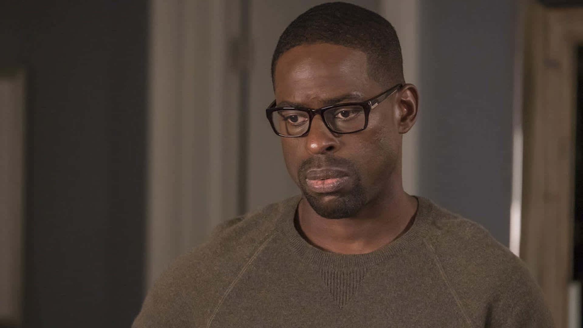 Sterling K Brown 桌布 壁紙 — 一名戴眼鏡、身穿棕色毛衣的男子在昏暗的室內環境中專注地向前凝視。