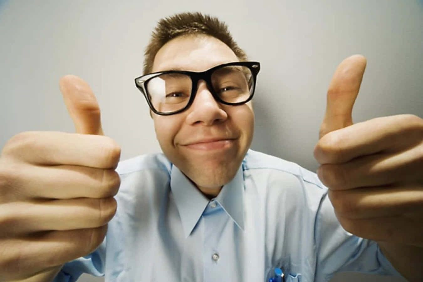 Nerd 桌布 壁紙 — 一名戴眼鏡、身穿淺藍色襯衫的男子，在純灰色背景前，露出燦爛笑容，熱情地豎起兩個大拇指。