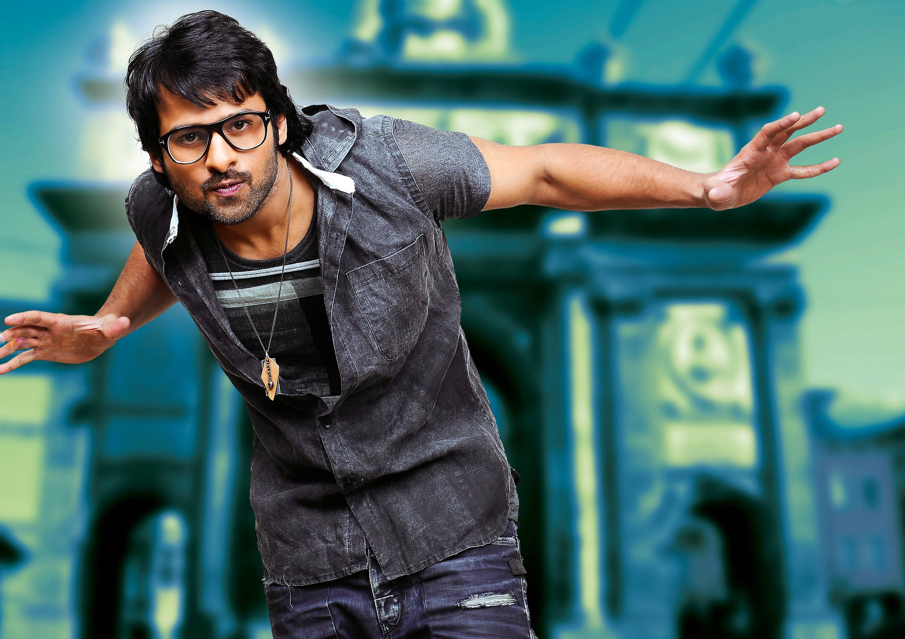 Prabhas HD 桌布 壁紙 — 一名戴眼鏡、身穿牛仔襯衫的男子，在模糊的藍綠色和綠色城市背景下擺出充滿動感的姿勢。