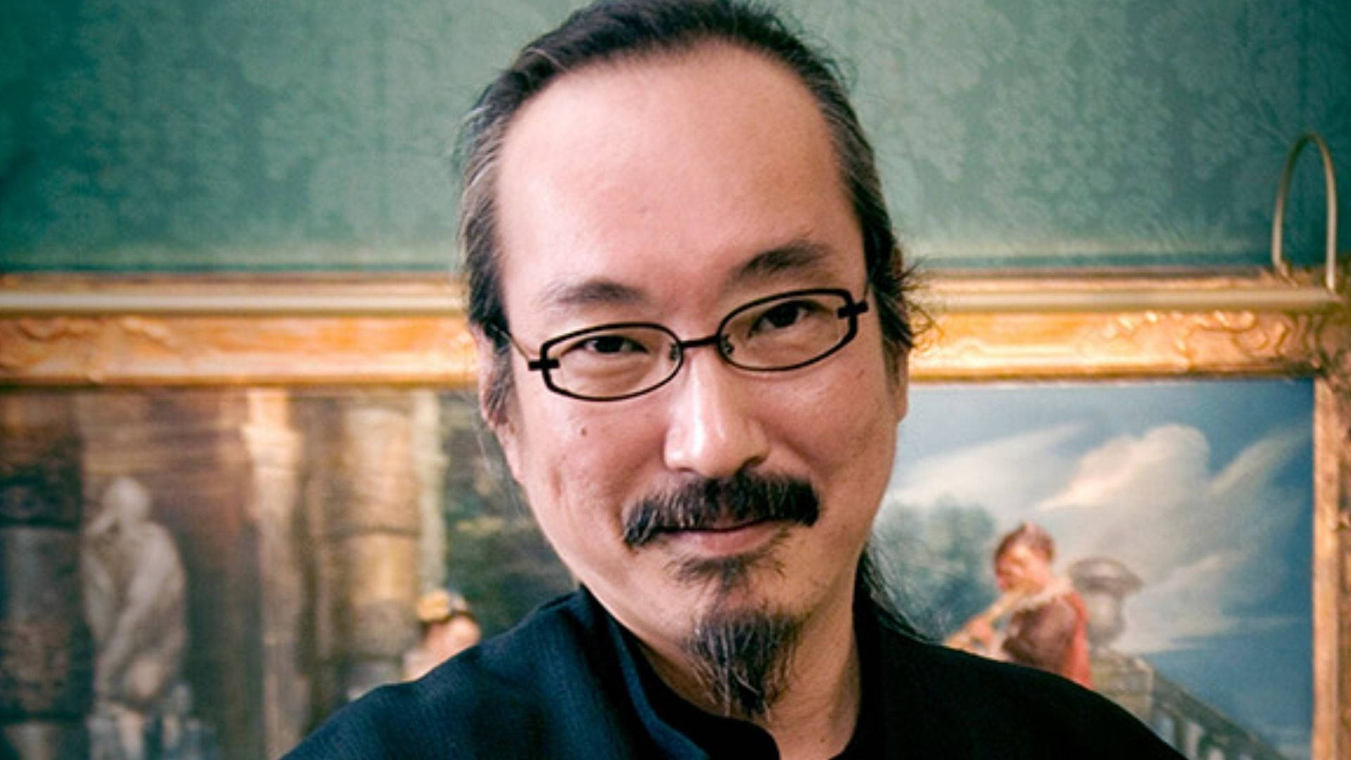 satoshi kon 桌布 壁紙 — 一名戴眼鏡及留鬍鬚的男子在青綠色牆上裝飾華麗的畫框前微笑。