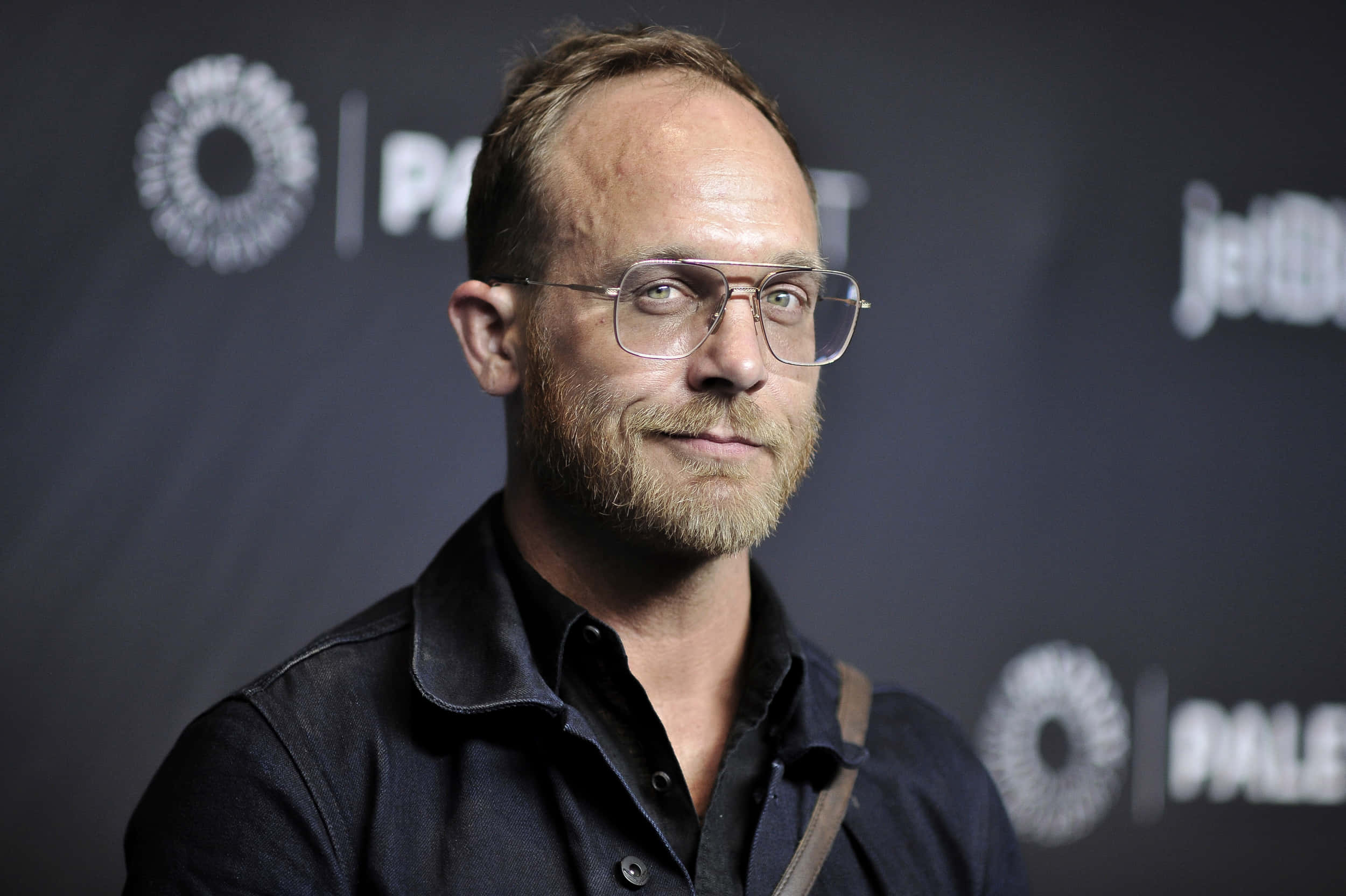 Ethan Embry 桌布 壁紙 — 一名戴眼鏡和鬍鬚的男子在深色品牌背景下微妙地微笑。
