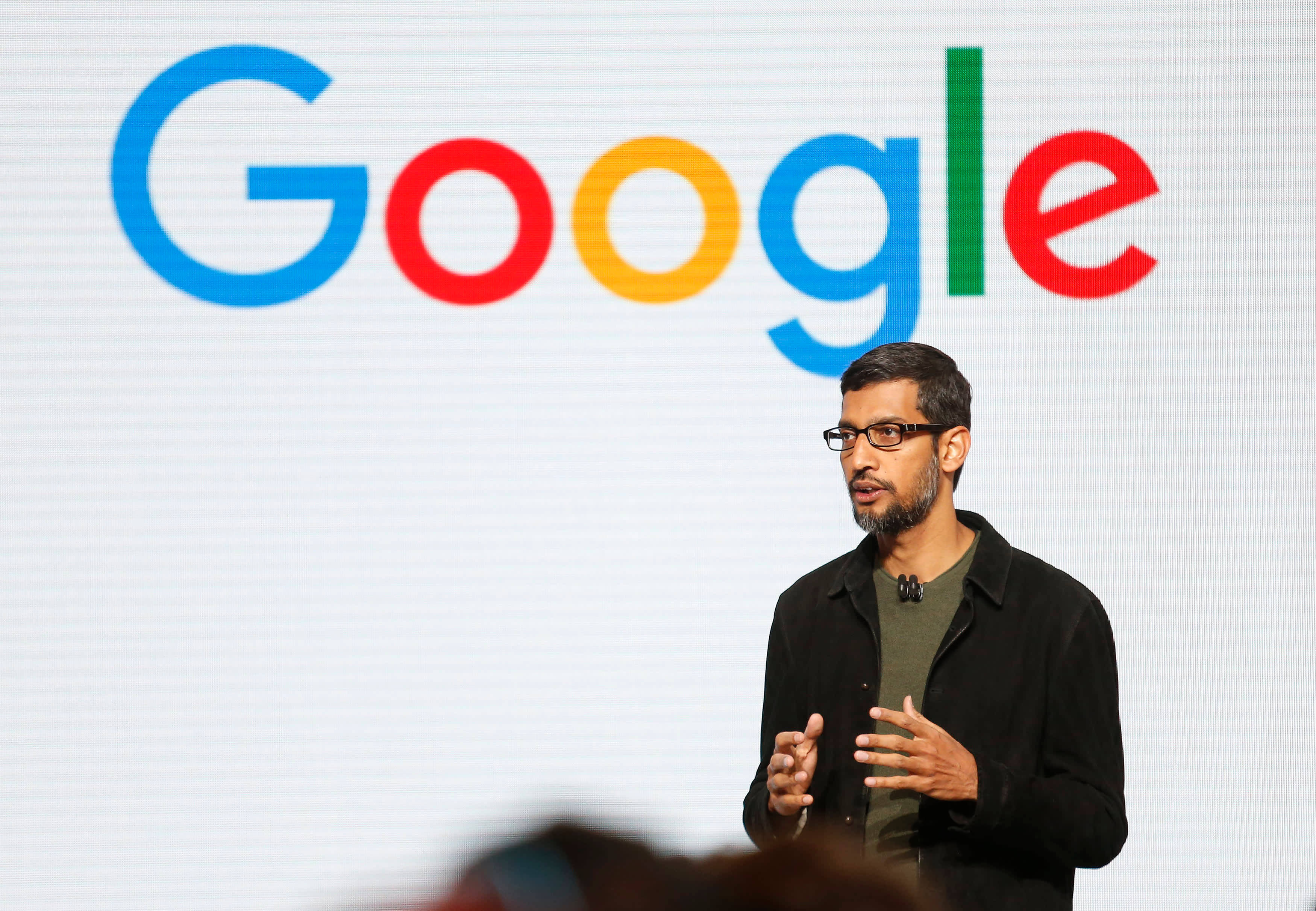 google 桌布 壁紙 — 一名戴眼鏡留鬍鬚的男子在舞台上，面前是大型彩色Google標誌螢幕，正在發言。