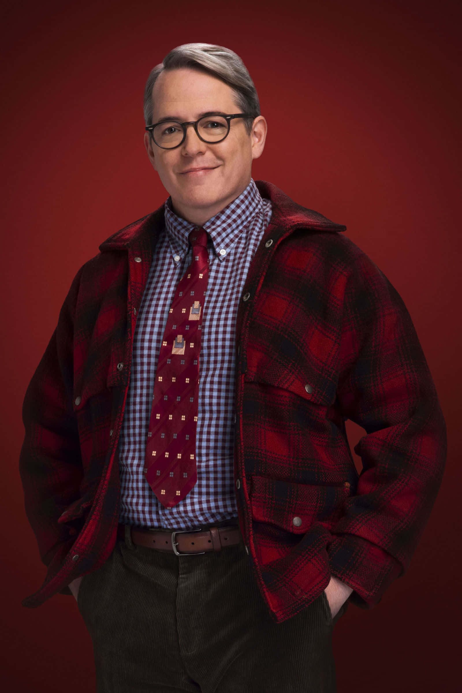 a christmas story 桌布 壁紙 — 一名戴眼鏡的男子，身穿紅色格紋外套，內搭格紋襯衫和圖案領帶，在深紅色背景前溫暖地微笑。