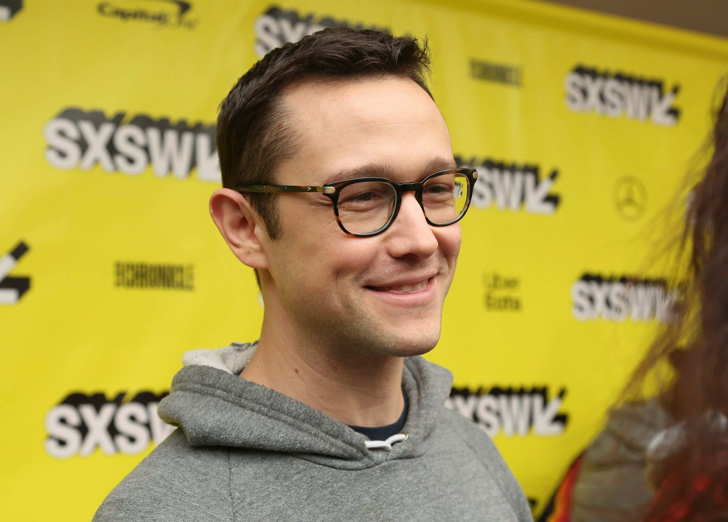 joseph gordon-levitt 桌布 壁紙 — 一名戴眼鏡的男子在明亮的黃色SXSW品牌背景前溫暖地微笑。