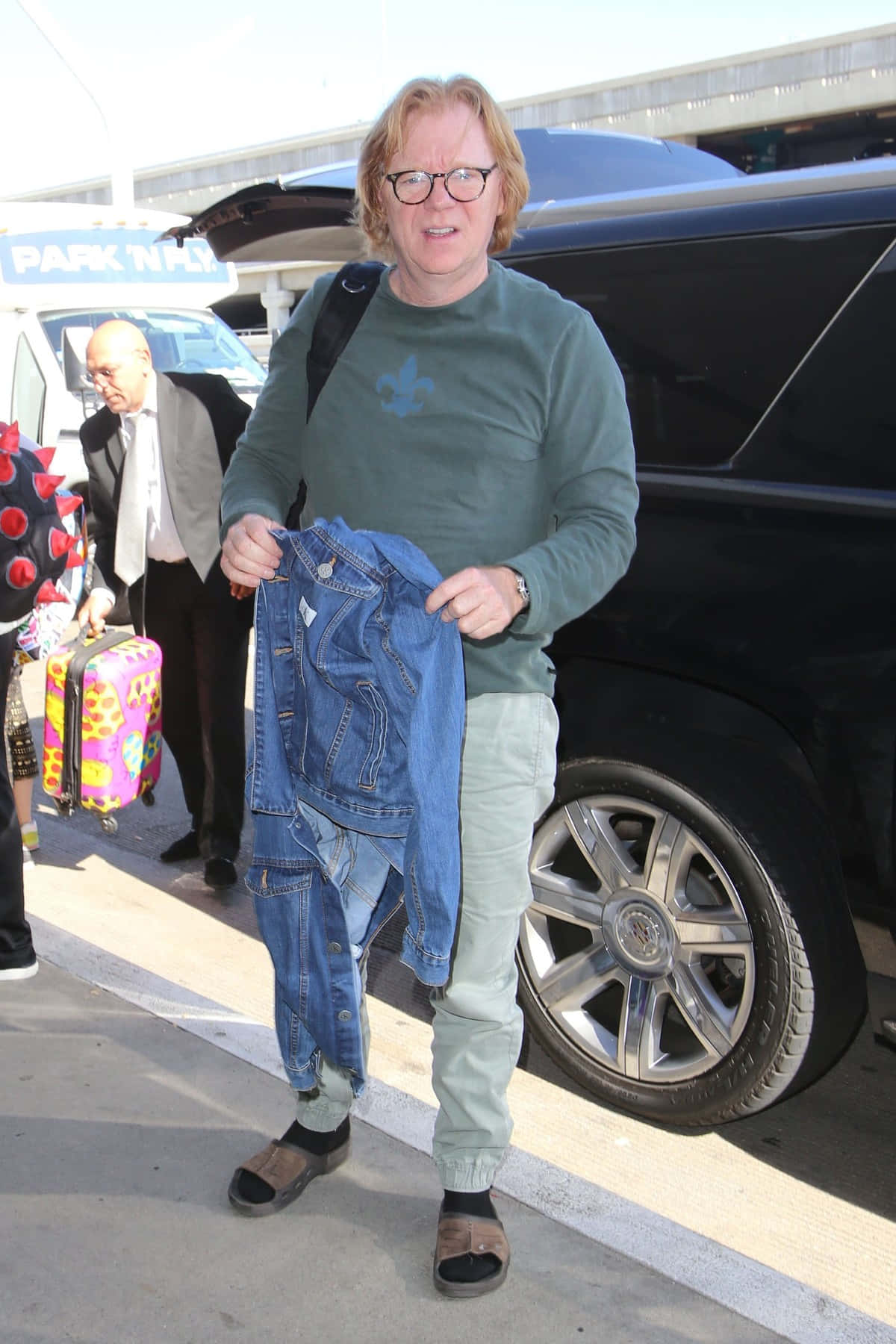 David Caruso 桌布 壁紙 — 一名戴眼鏡的男子在機場停車區站在一輛黑色SUV旁邊，舉起一件牛仔外套。