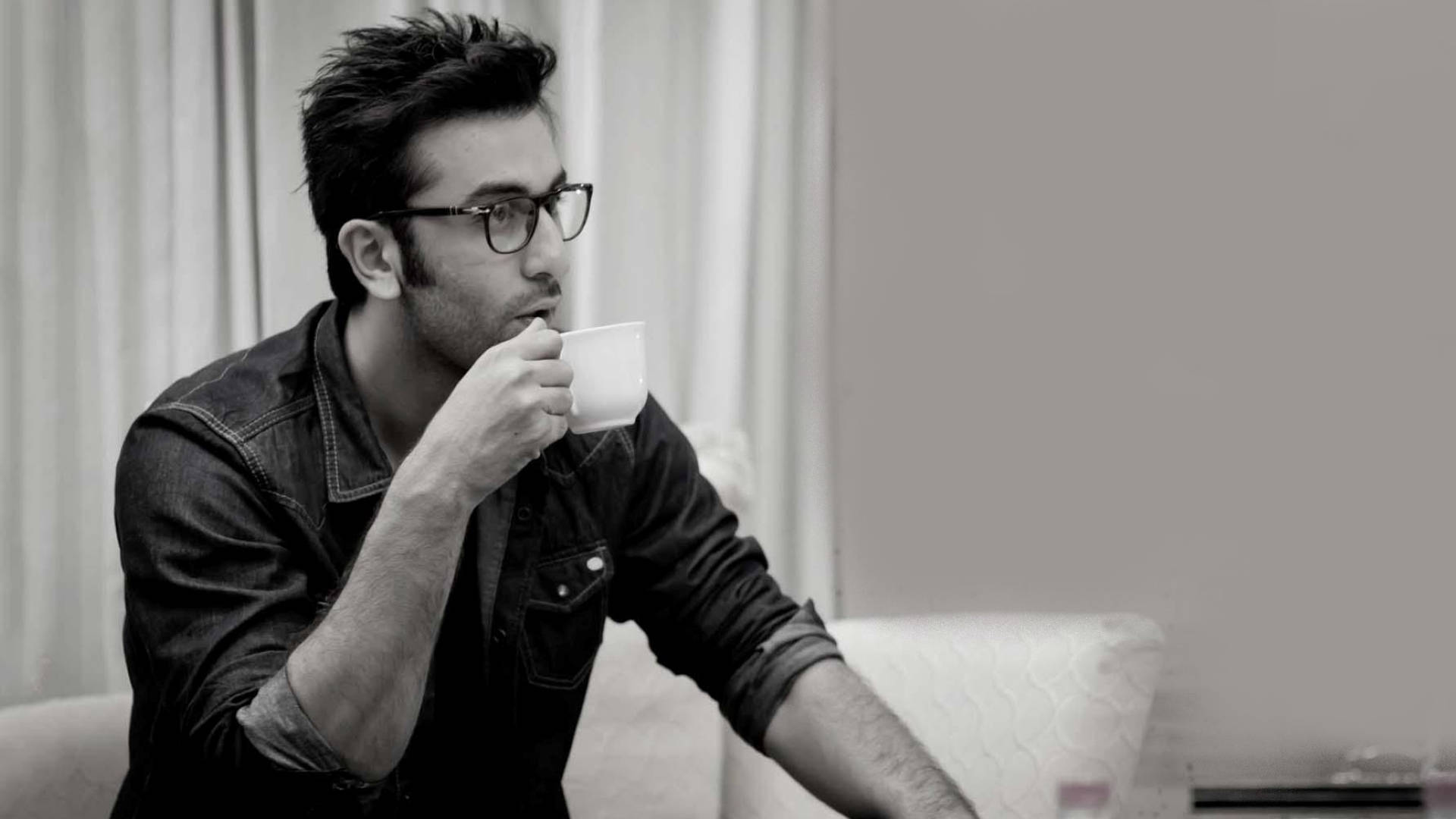 Ranbir Kapoor 桌布 壁紙 — 一名戴眼鏡的男子坐在沙發上，從白色馬克杯啜飲，身處柔和燈光的單色室內。
