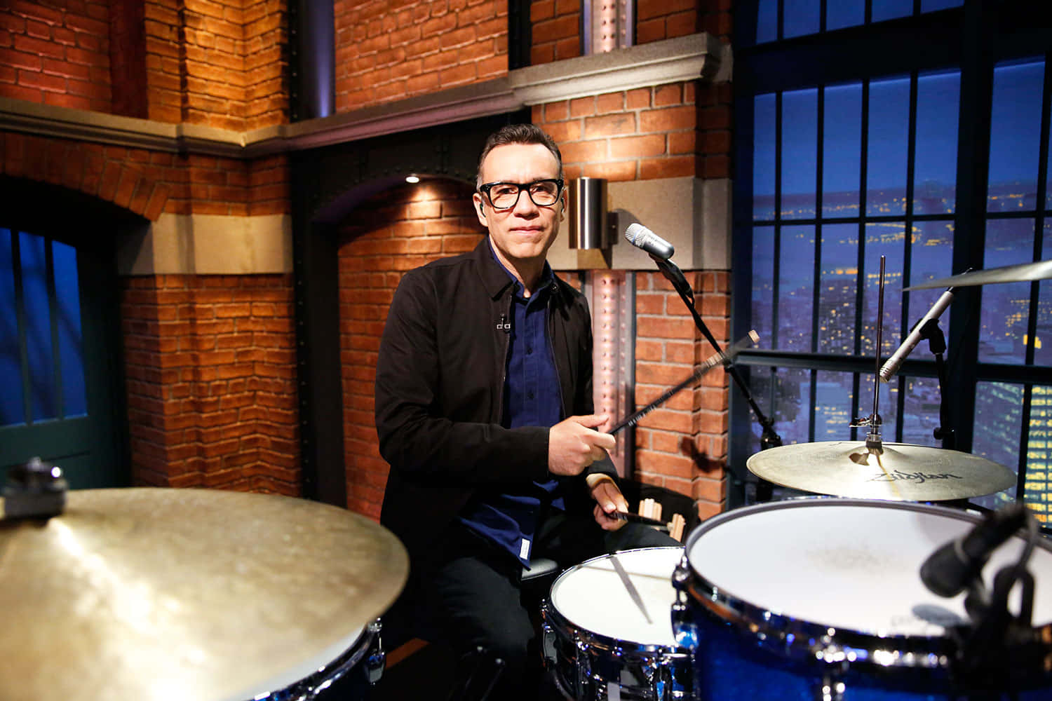 fred armisen 桌布 壁紙 — 一名戴眼鏡的男子坐在磚牆錄音室的鼓組後面，俯瞰夜晚發光的城市景觀。