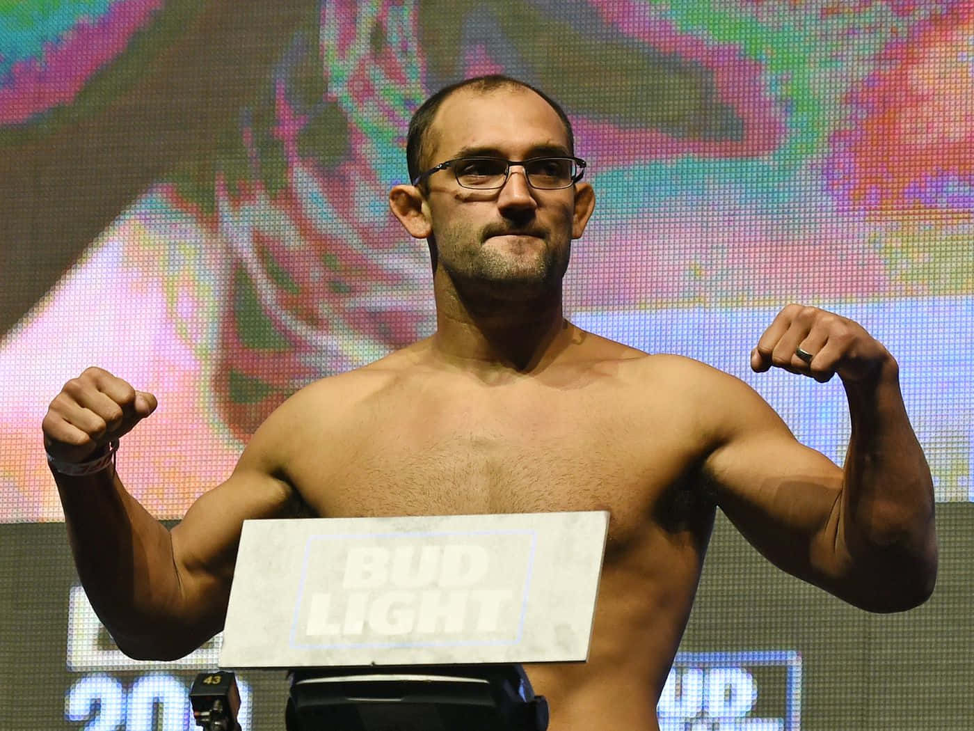 UFC 桌布 壁紙 — 一名戴眼鏡的肌肉發達拳手在色彩繽紛的LED螢幕背景的稱重活動中，在磅上彎曲雙臂。