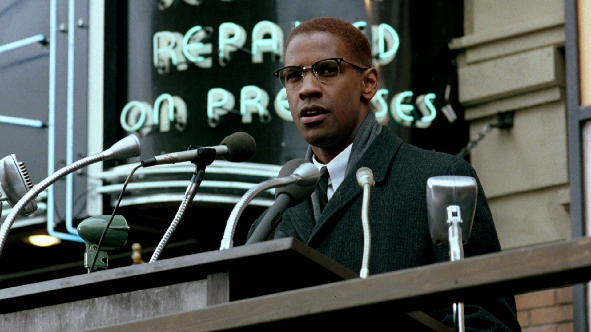 Denzel Washington 桌布 壁紙 — 一名戴眼鏡穿西裝的男子在寫有「REPAID ON PROMISES」的霓虹燈牌下，於設有多個麥克風的講台上發言。