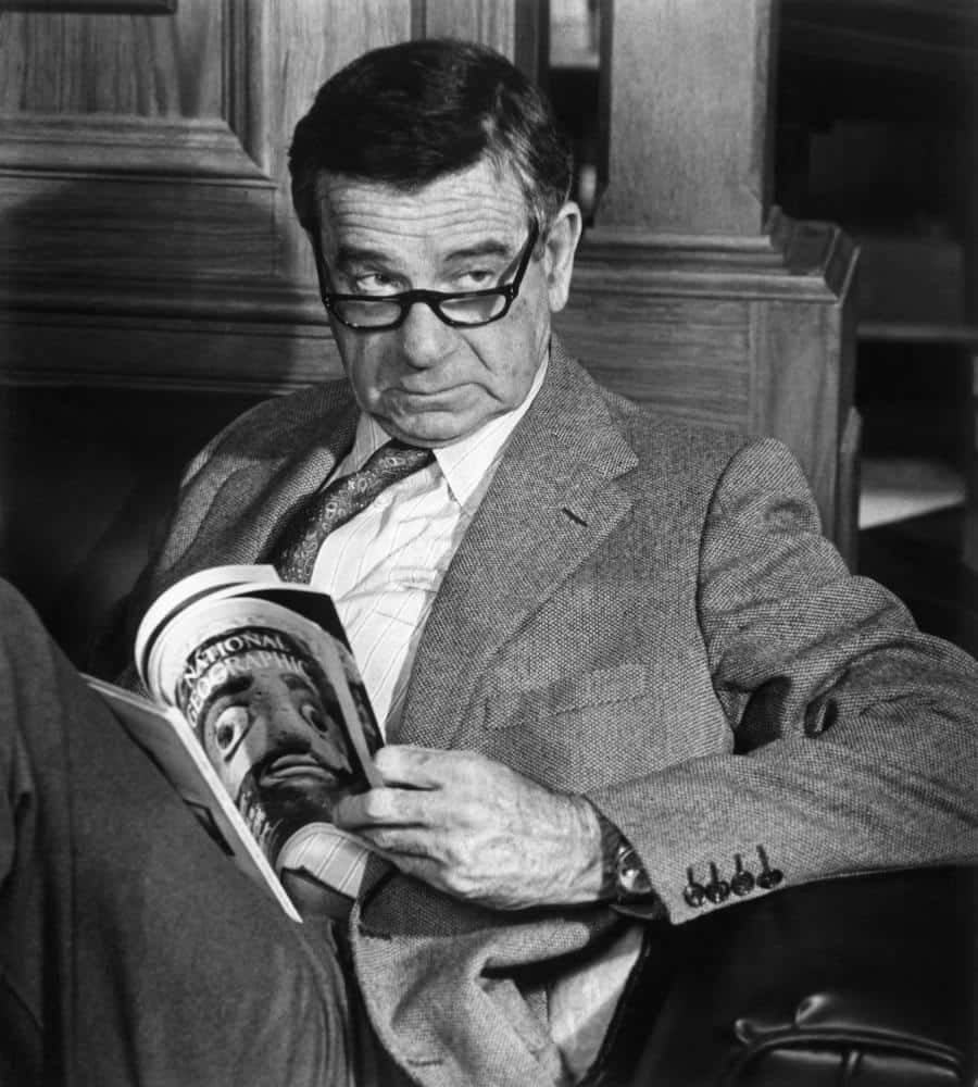 Walter Matthau 桌布 壁紙 — 一名戴眼鏡穿西裝的男子坐在木椅上閱讀《國家地理》雜誌。