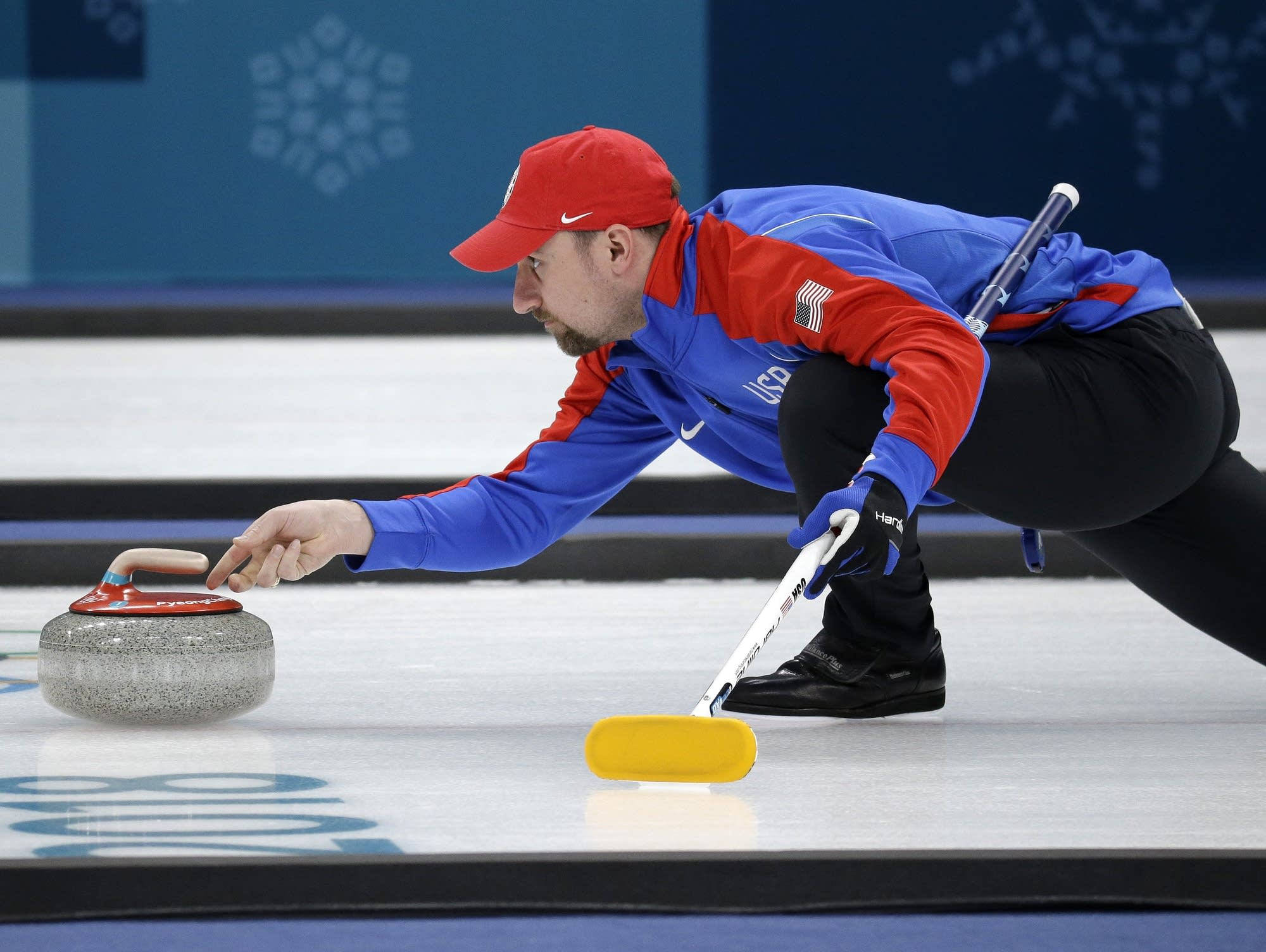 Olympics 桌布 壁紙 — 一名戴紅帽、穿藍色運動衫的男性冰壺運動員在比賽期間在冰面上推著一塊花崗岩石頭。
