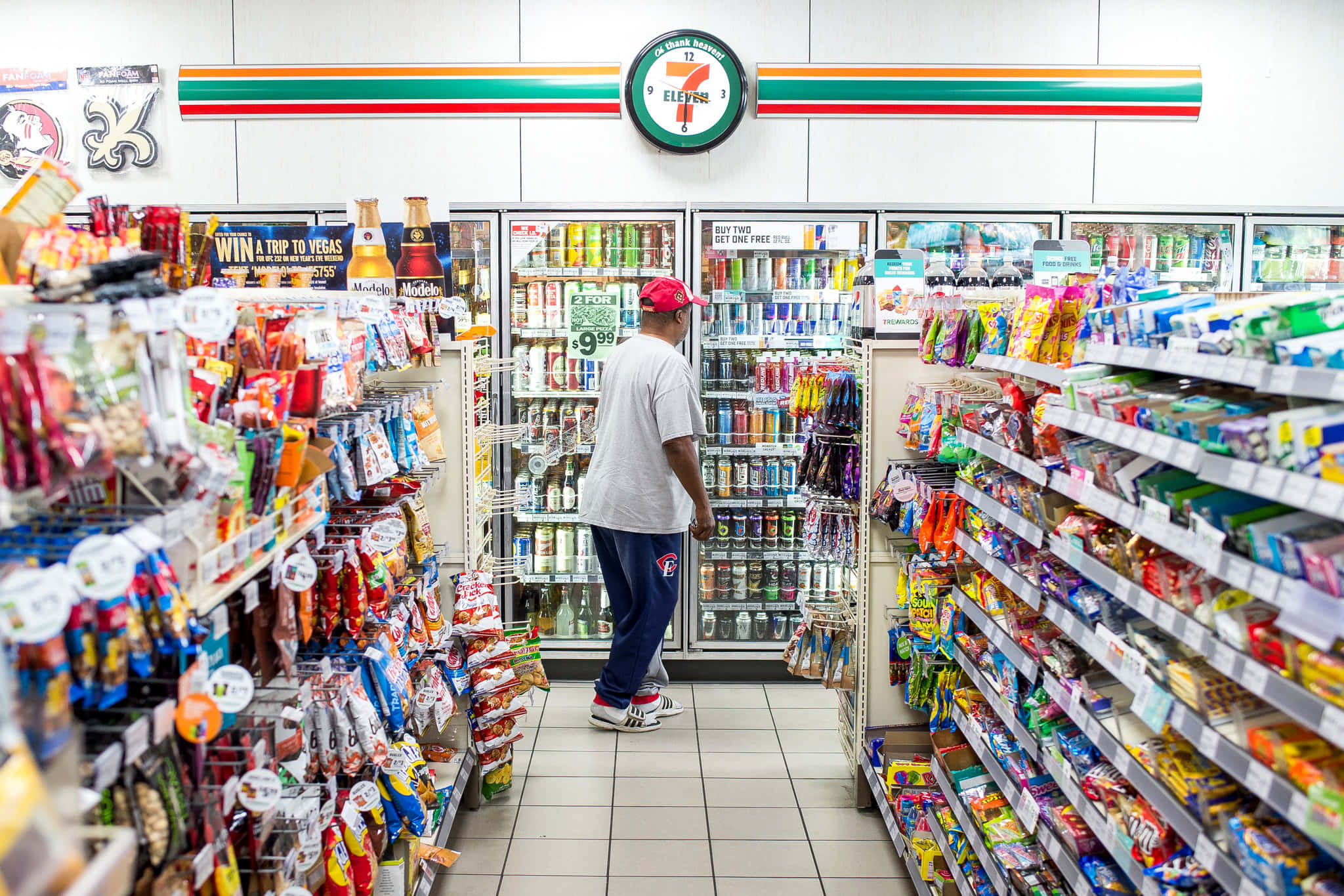 7-ELEVEN 桌布 壁紙 — 一名戴紅帽的男子在燈光明亮的7-Eleven通道瀏覽，通道上擺滿了色彩繽紛的零食和飲料。