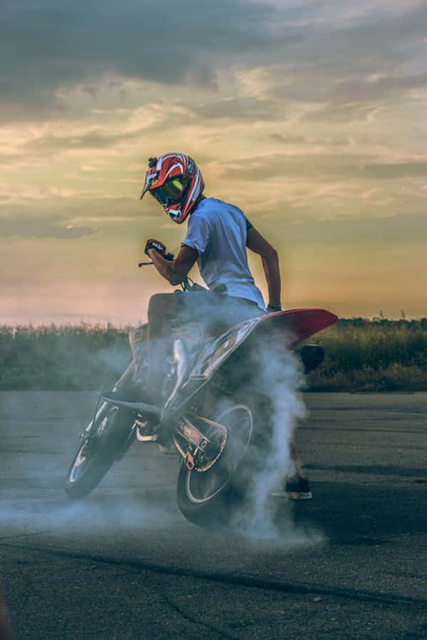桌布 壁紙 — 一名戴紅色安全帽的摩托車手在日落時分的柏油路上進行充滿煙霧的燒胎。