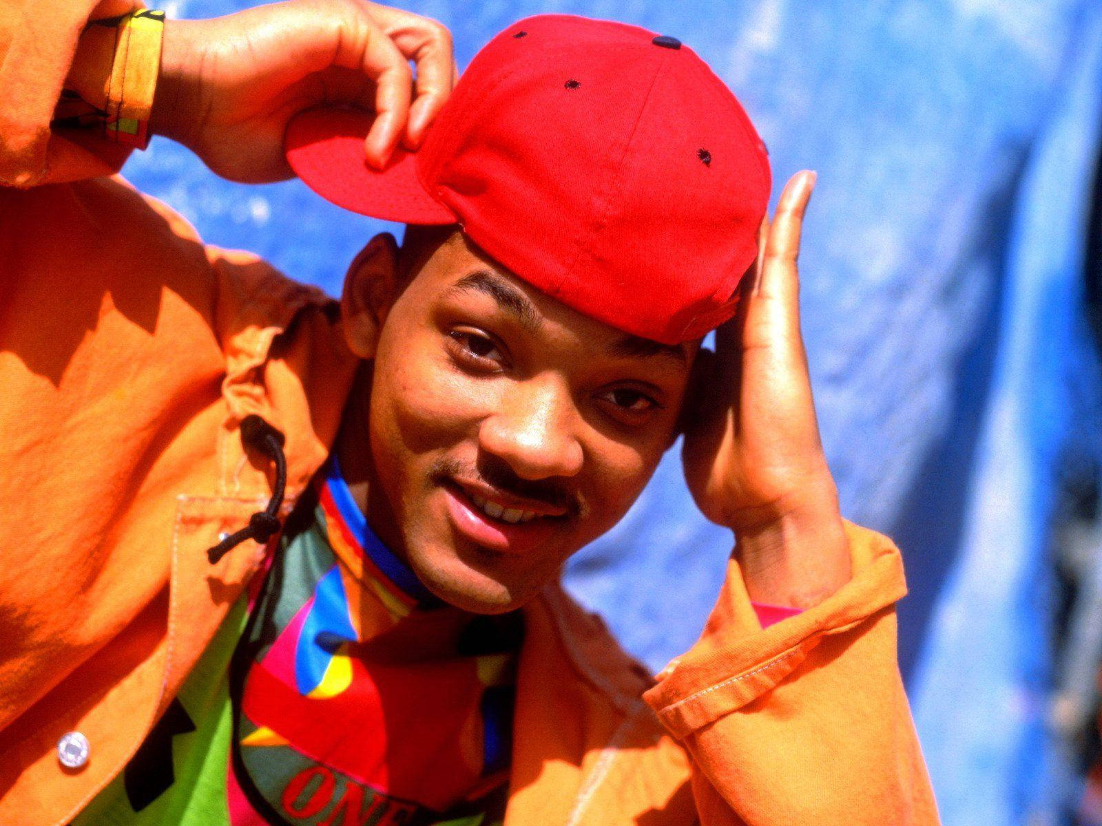 fresh prince 桌布 壁紙 — 一名戴紅色帽、穿橙色外套的年輕男子在鮮豔的藍色背景前微笑著調整帽子。