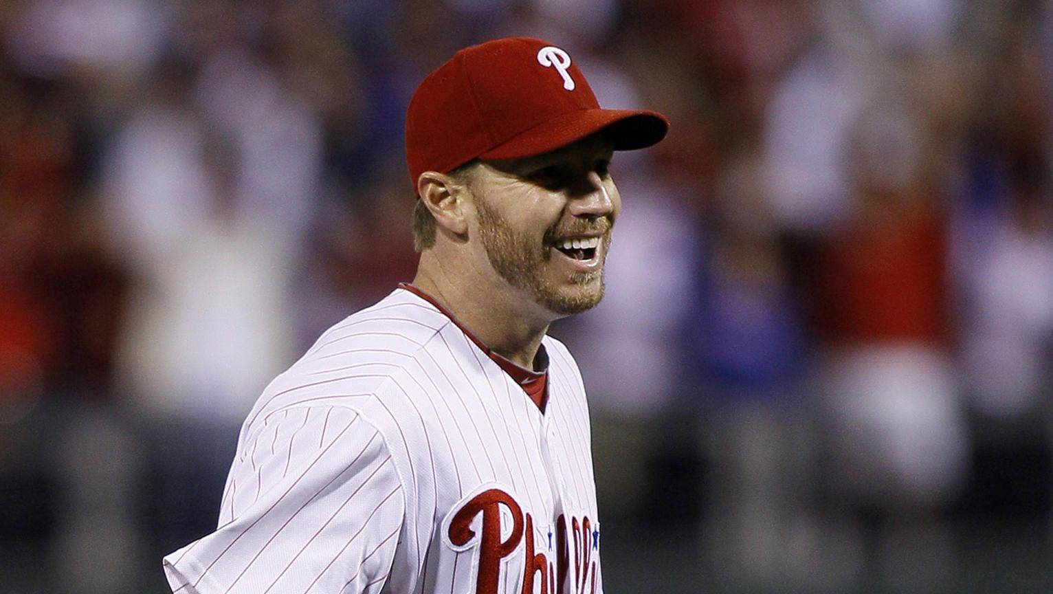 roy halladay 桌布 壁紙 — 一名戴紅色帽、身穿白色條紋球衣的棒球運動員在球場燈光下的夜間比賽中開懷大笑。