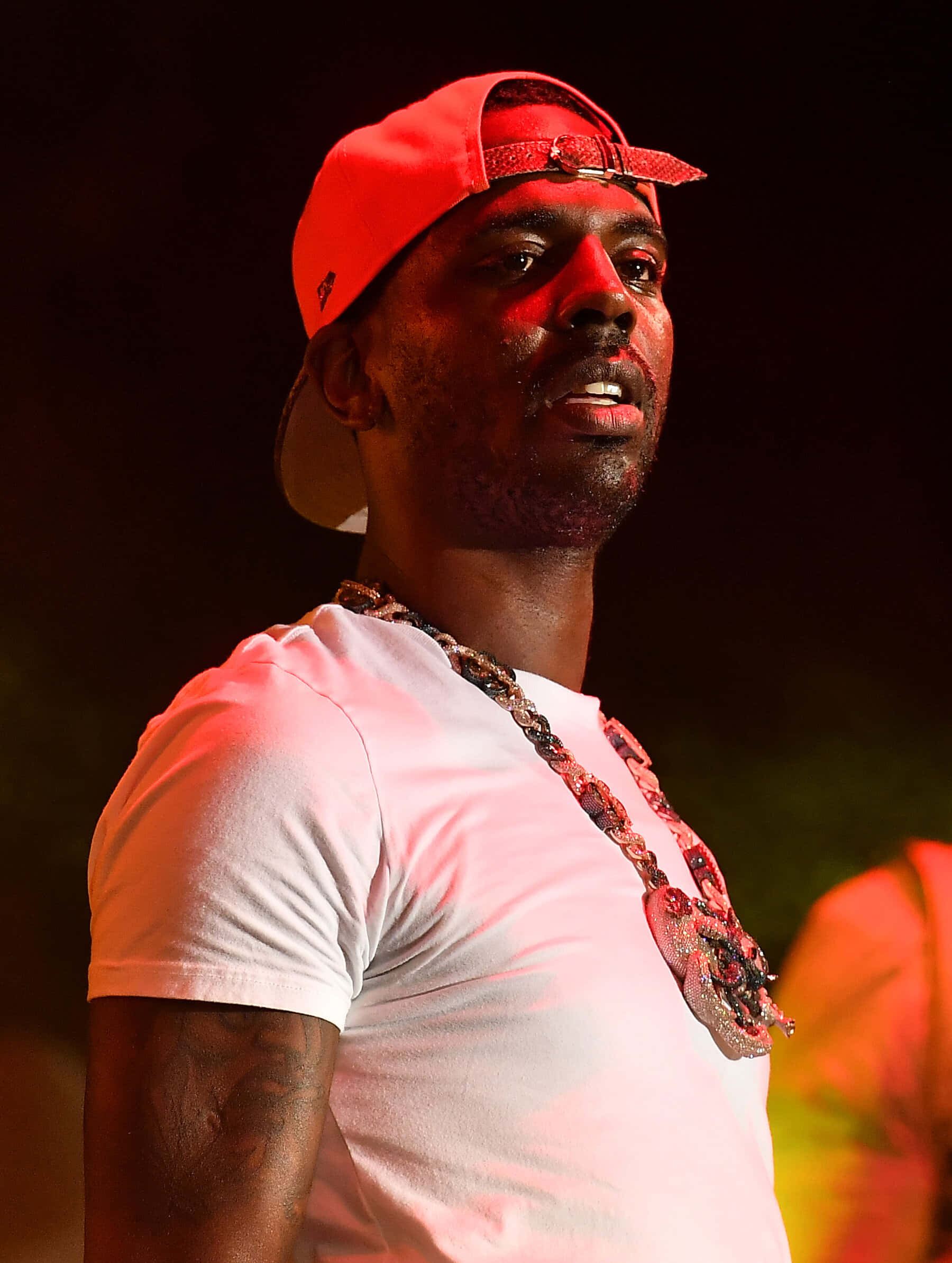 Young Dolph 桌布 壁紙 — 一名戴紅色帽子、穿白色T恤的男人，在戲劇性的紅色舞台燈光下表演，戴著一條粗鏈條項鍊。