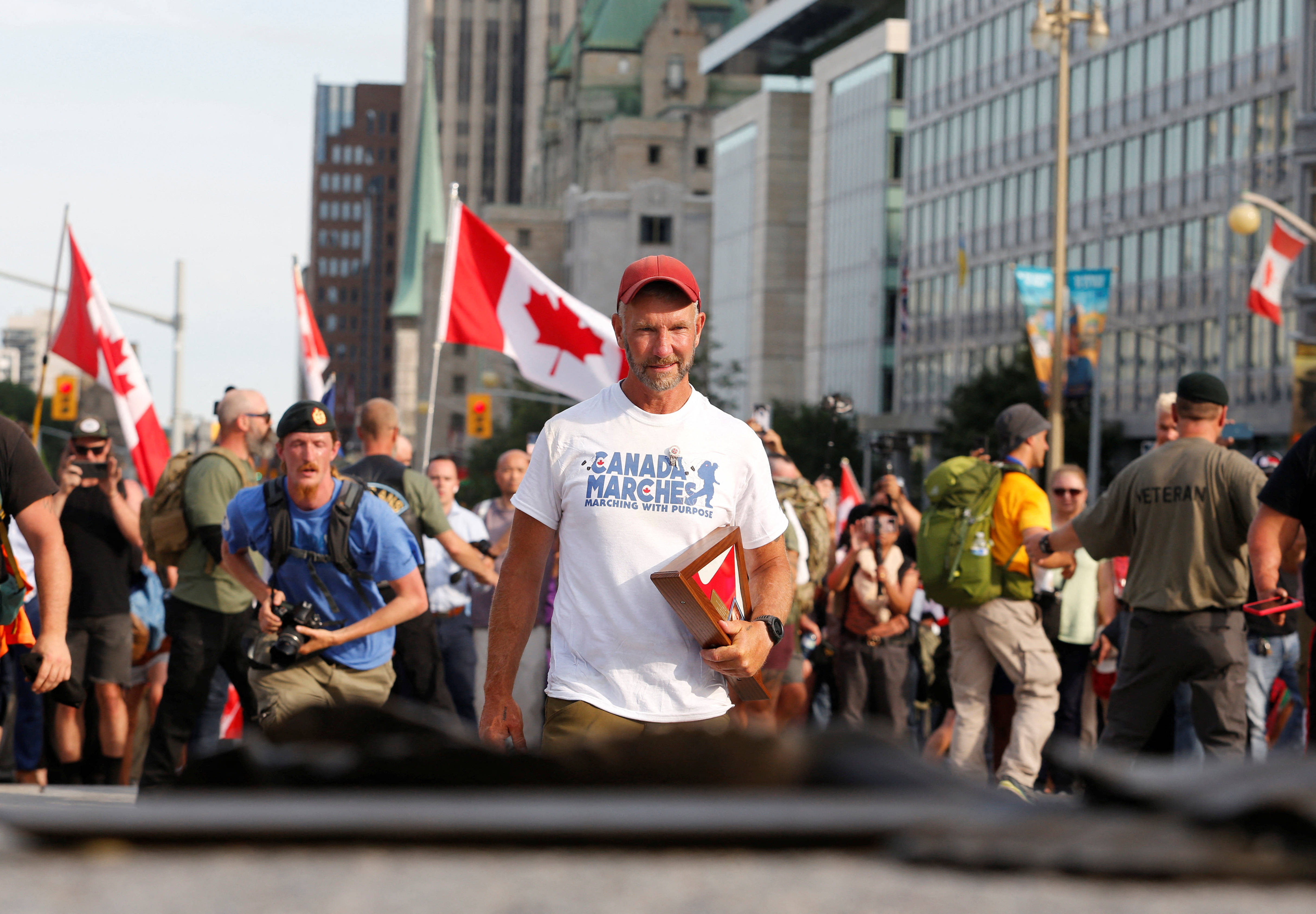 Flag 桌布 壁紙 — 一名戴紅色帽子和身穿「Canada Marches」T恤的男子，在日間抗議活動中手持裱框照片向前走，背景有加拿大國旗和城市建築。