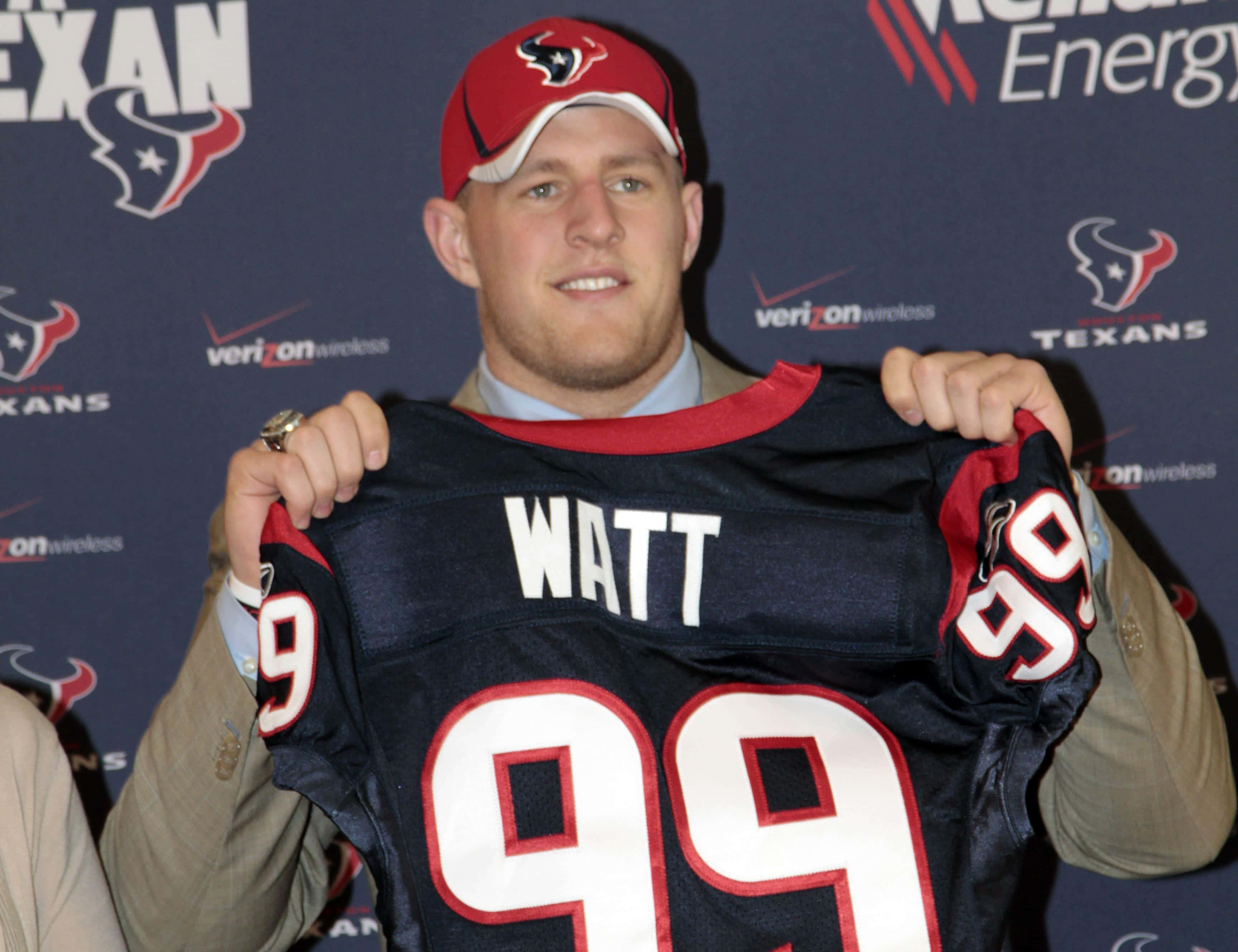 Jj Watt 桌布 壁紙 — 一名戴紅色帽子的微笑男子，在品牌背景前舉起一件印有WATT名字和99號碼的休士頓德州人球衣。