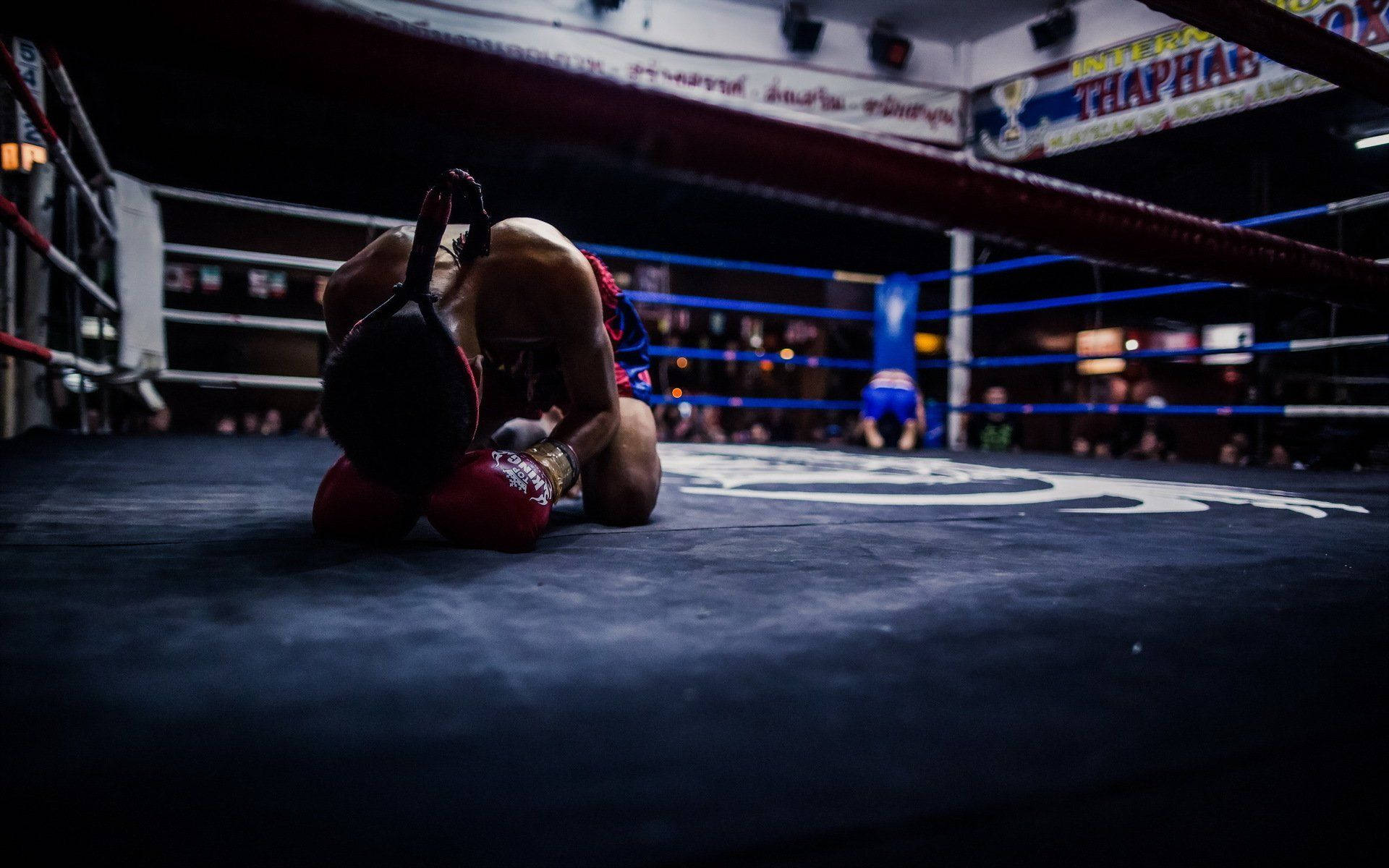 Boxing 桌布 壁紙 — 一名戴紅色手套的拳擊手在黑暗的擂台上跪地失敗，另一名拳手在背景中勝利站立。