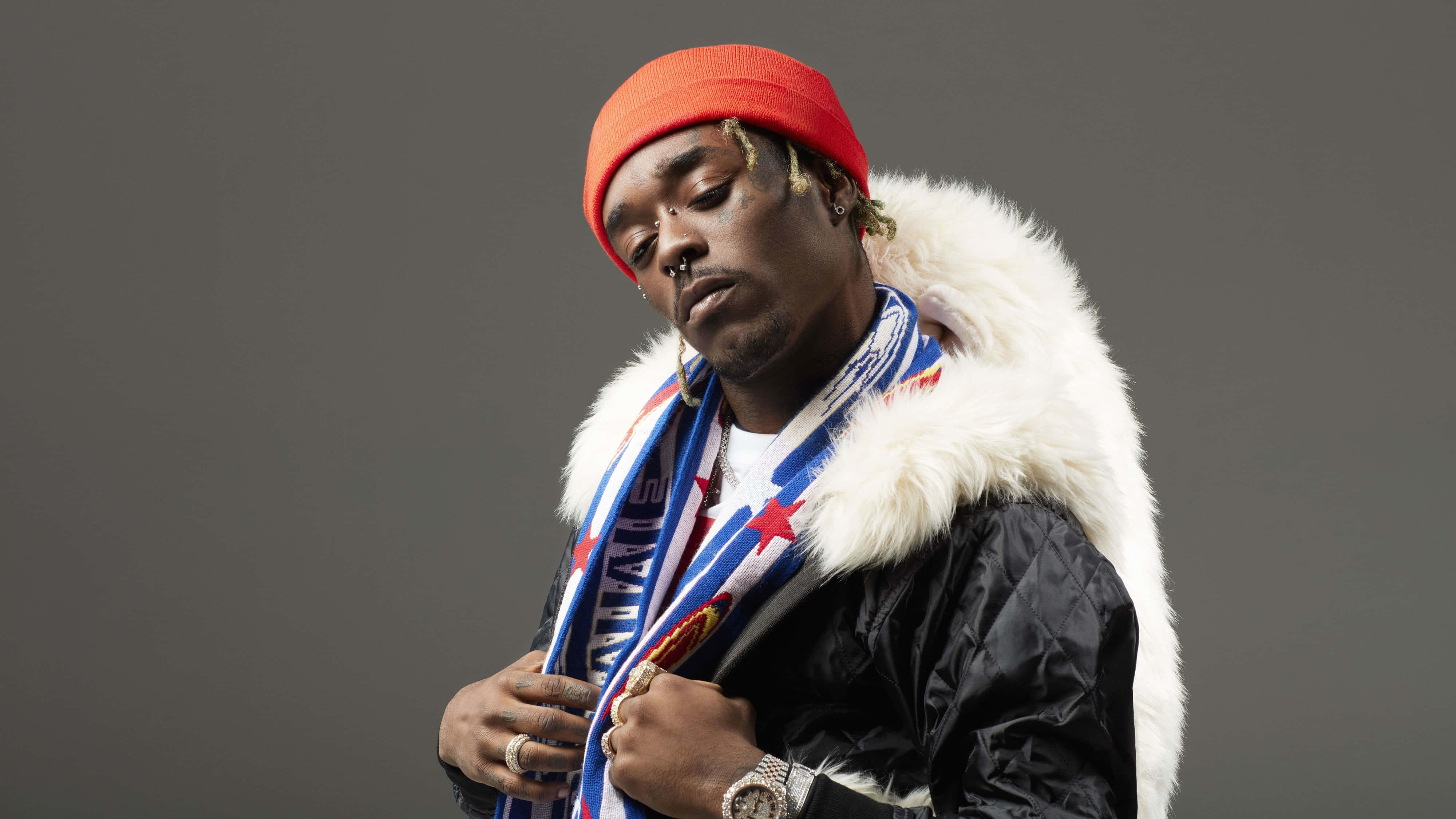 Lil Uzi Vert 桌布 壁紙 — 一名戴紅色毛帽、穿毛領外套的男子，在純灰色背景前閉眼擺姿勢。