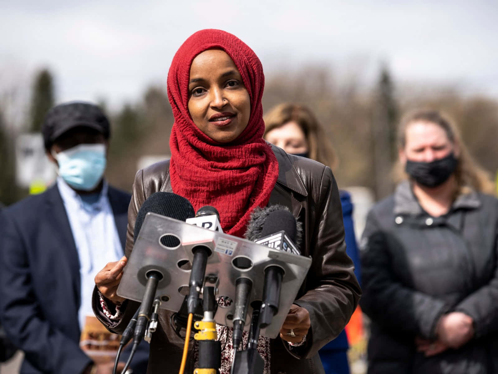 Hijab 桌布 壁紙 — 一名戴紅色頭巾的女性在戶外記者會上發言，身後有麥克風和戴口罩的人。