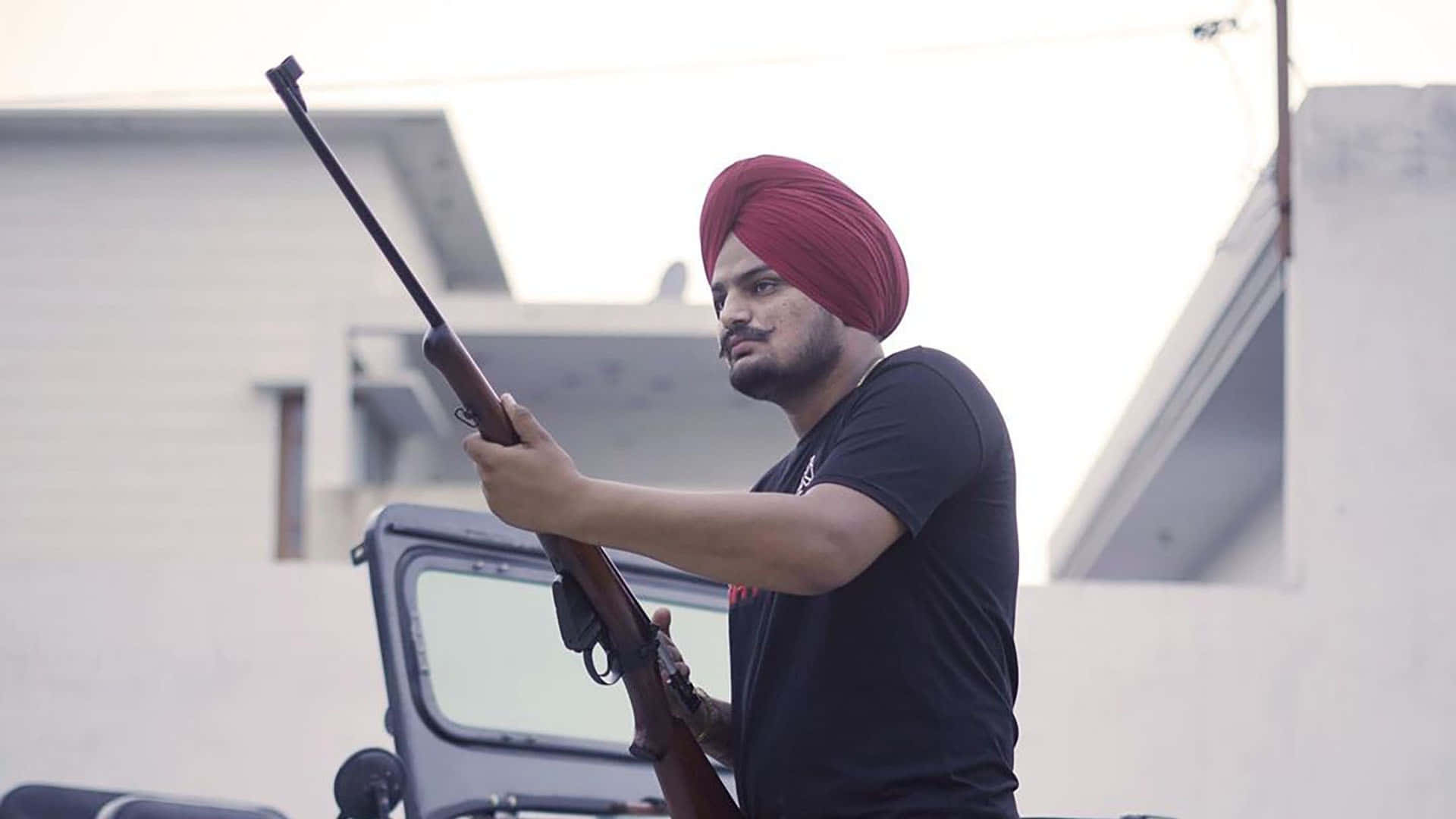 戴紅色頭巾的男子在建築物附近戶外持步槍 — Sidhu Moose Wala 壁紙
