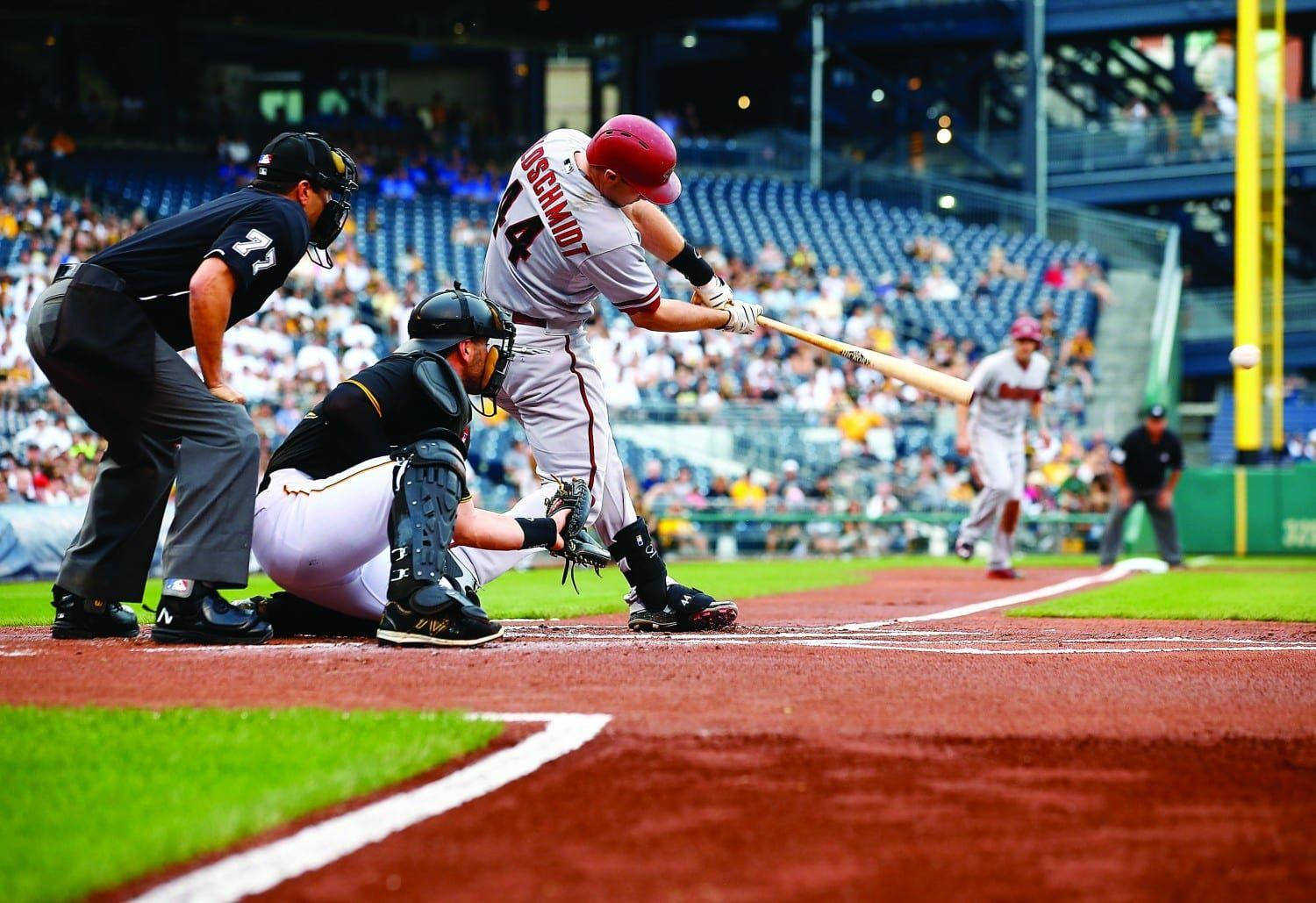 Paul Goldschmidt 桌布 壁紙 — 一名戴紅色頭盔的棒球打者在陽光明媚的球場上揮棒擊球，捕手和裁判蹲伏在本壘板後方。