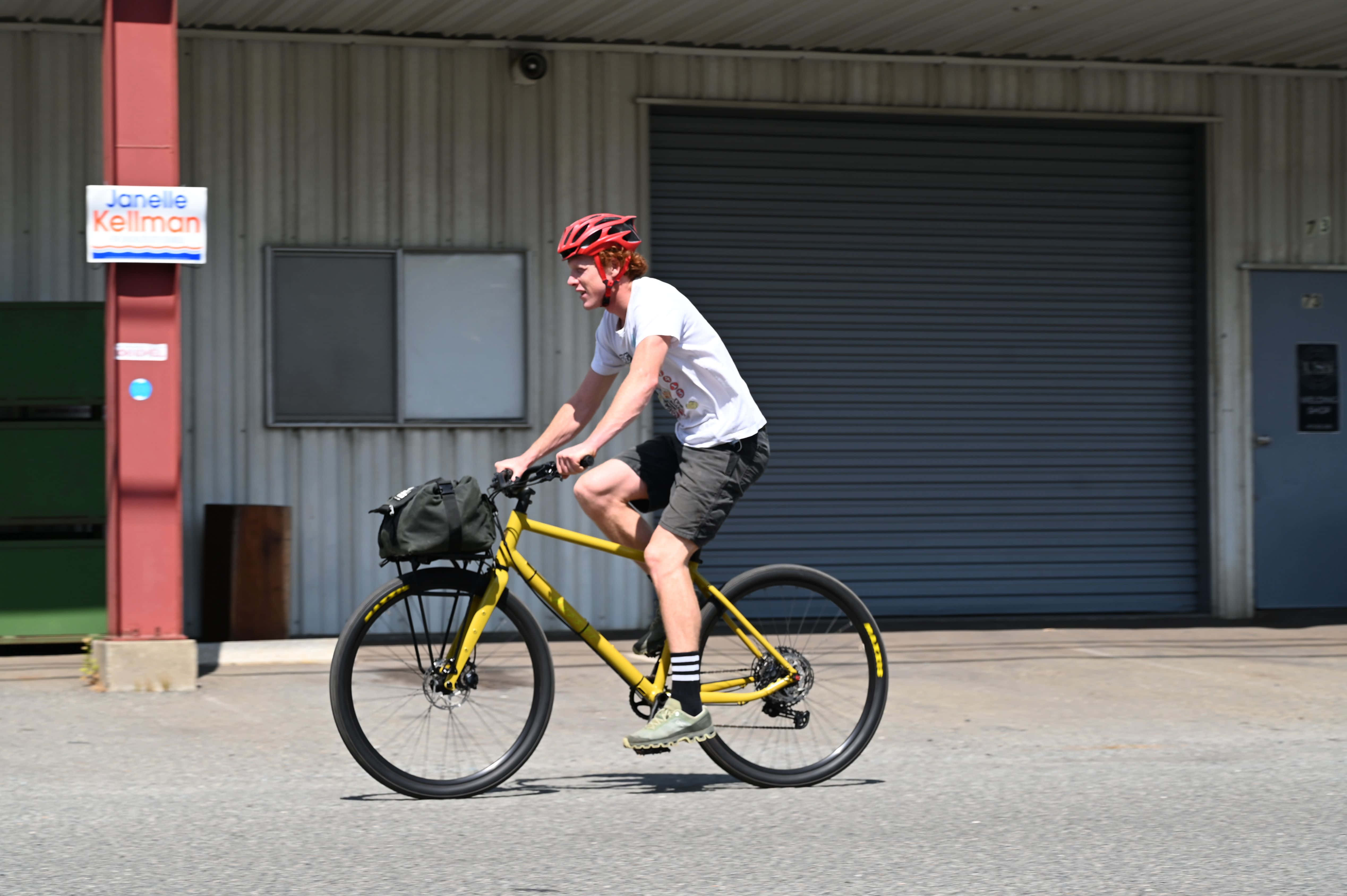 Bicycle 桌布 壁紙 — 一名戴紅色頭盔的自行車騎士在晴朗日光下，騎著亮黃色自行車，經過一座波紋金屬建築前的鋪砌空地。