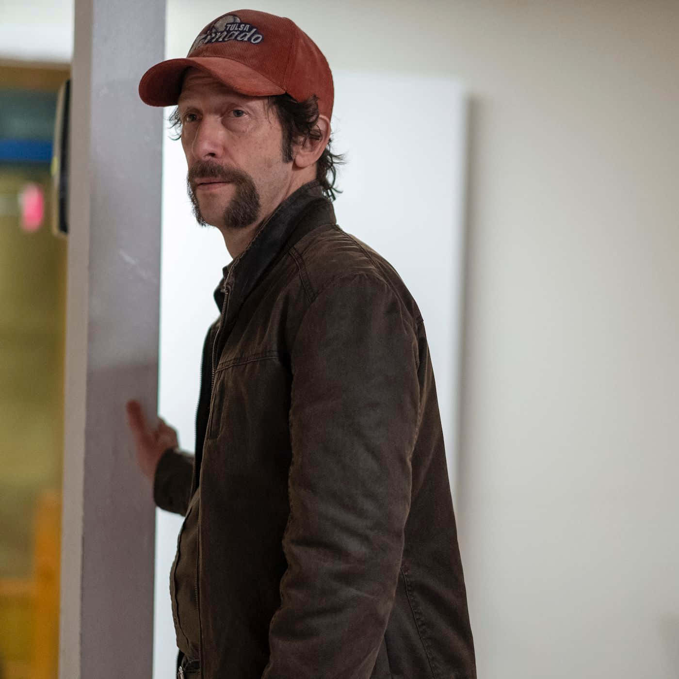 Tim Blake Nelson 桌布 壁紙 — 一名戴紅色鴨舌帽、身穿棕色外套的男子，表情嚴肅地靠在牆上。