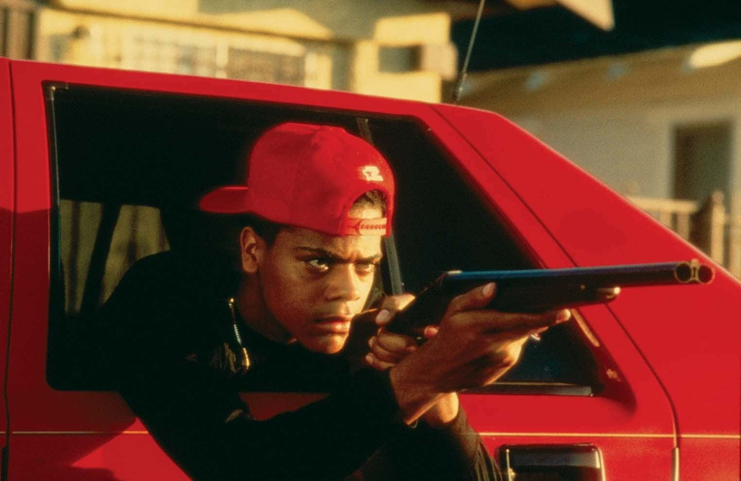 Boyz N The Hood 桌布 壁紙 — 一名戴紅色鴨舌帽的年輕人在溫暖的金色光線下，從紅色貨車的車窗瞄準一把霰彈槍。