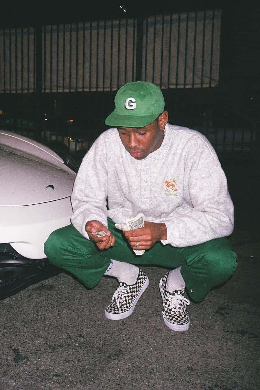 Rappers 桌布 壁紙 — 一名戴綠色帽及穿運動褲的男子蹲在白色車旁，在夜間城市燈光下數現金。