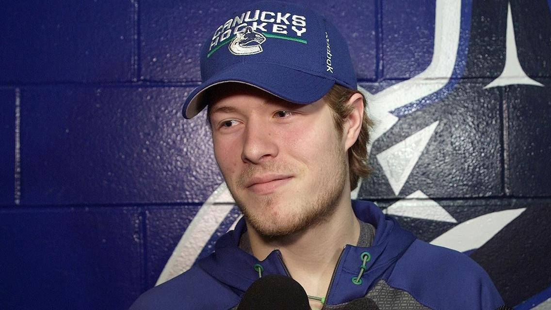 運動 桌布 壁紙 — 一名戴著 Canucks 帽子和藍色連帽衫的曲棍球運動員，在帶有球隊標誌的藍色磚牆前對著麥克風說話，微微一笑。