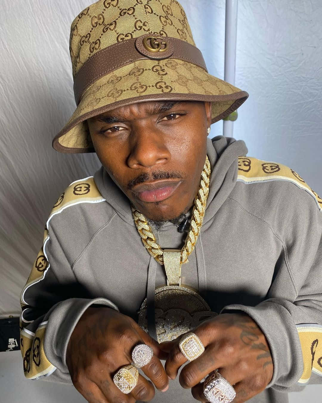 Dababy 桌布 壁紙 — 一名戴著Gucci漁夫帽和連帽衫的男子，展示著超大鑽石戒指和粗金鏈，眼神銳利。