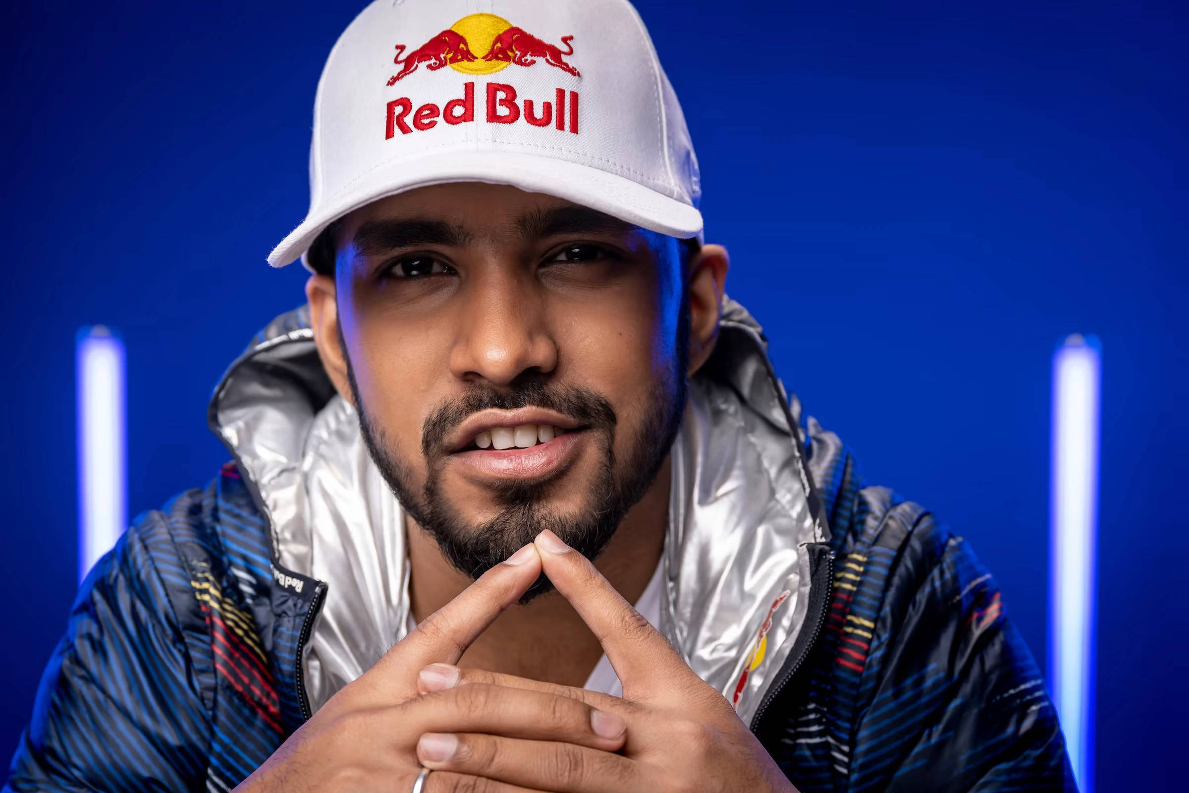 Red Bull 桌布 壁紙 — 一名戴著Red Bull帽子和閃亮外套的男子，在帶有發光垂直燈光的鮮豔藍色背景前，雙手靠近下巴擺姿勢。