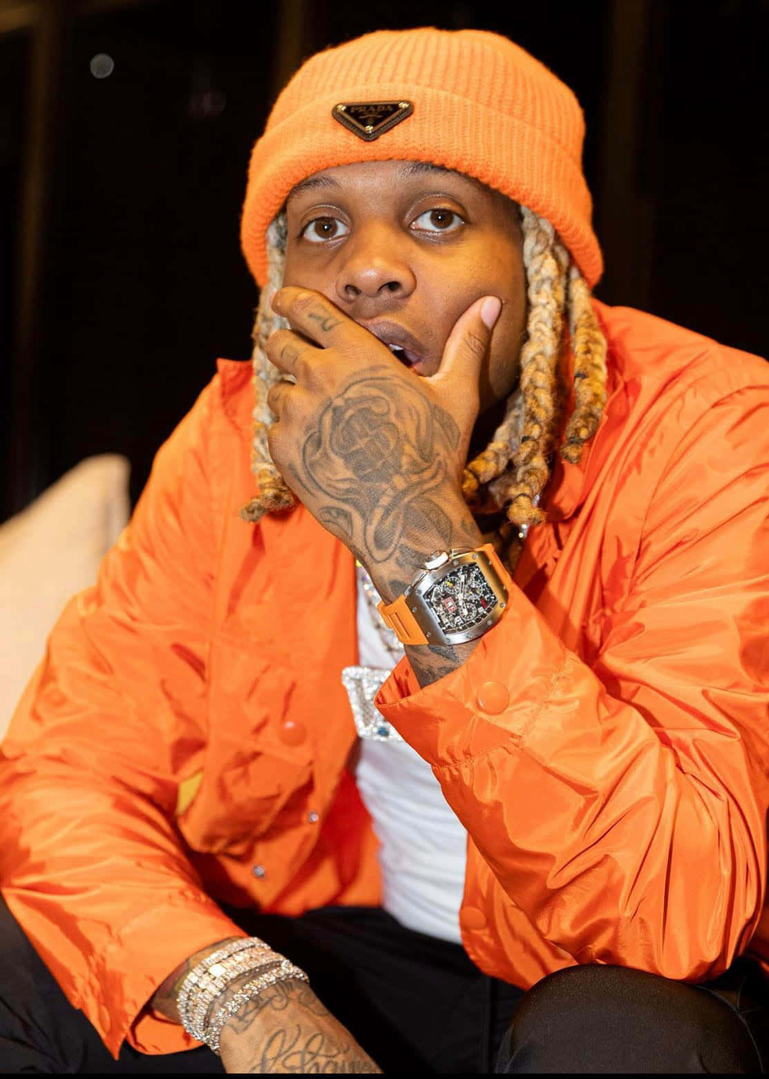 Lil Durk 桌布 壁紙 — 一名戴著亮橙色毛帽和相配外套的男子，將有刺青的手放在臉旁，佩戴奢華珠寶擺姿勢。